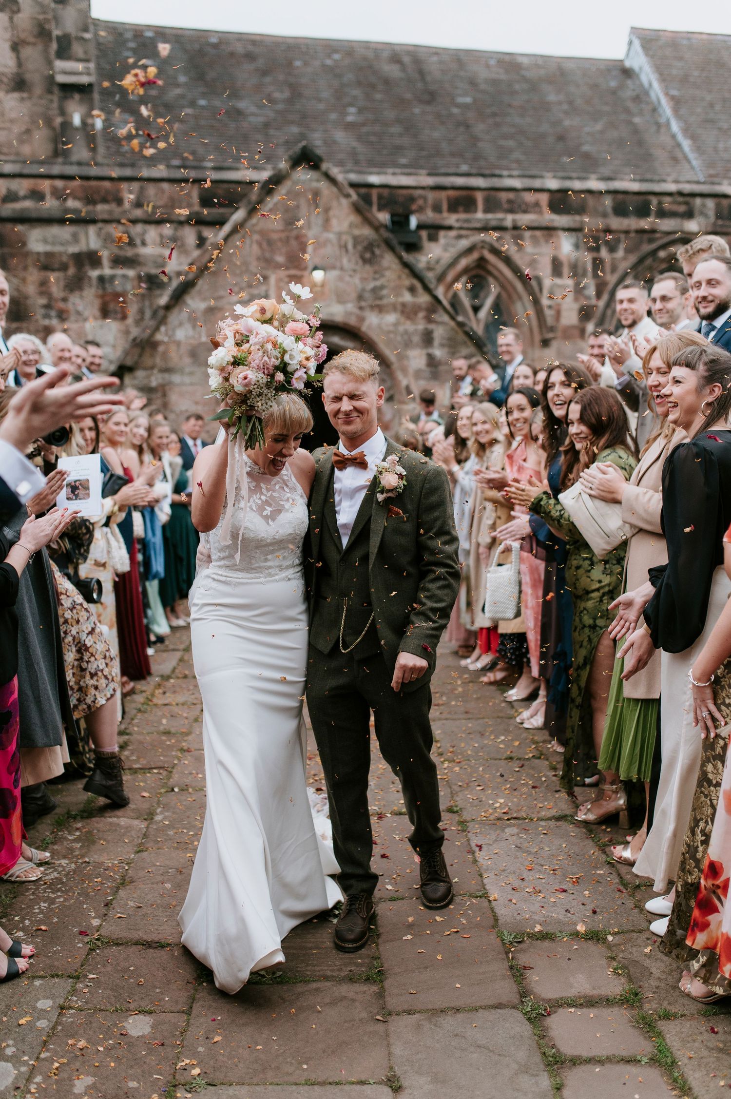 Wedding guests throw flower petals as newlyweds walk through a stone courtyard during their celebration exit.