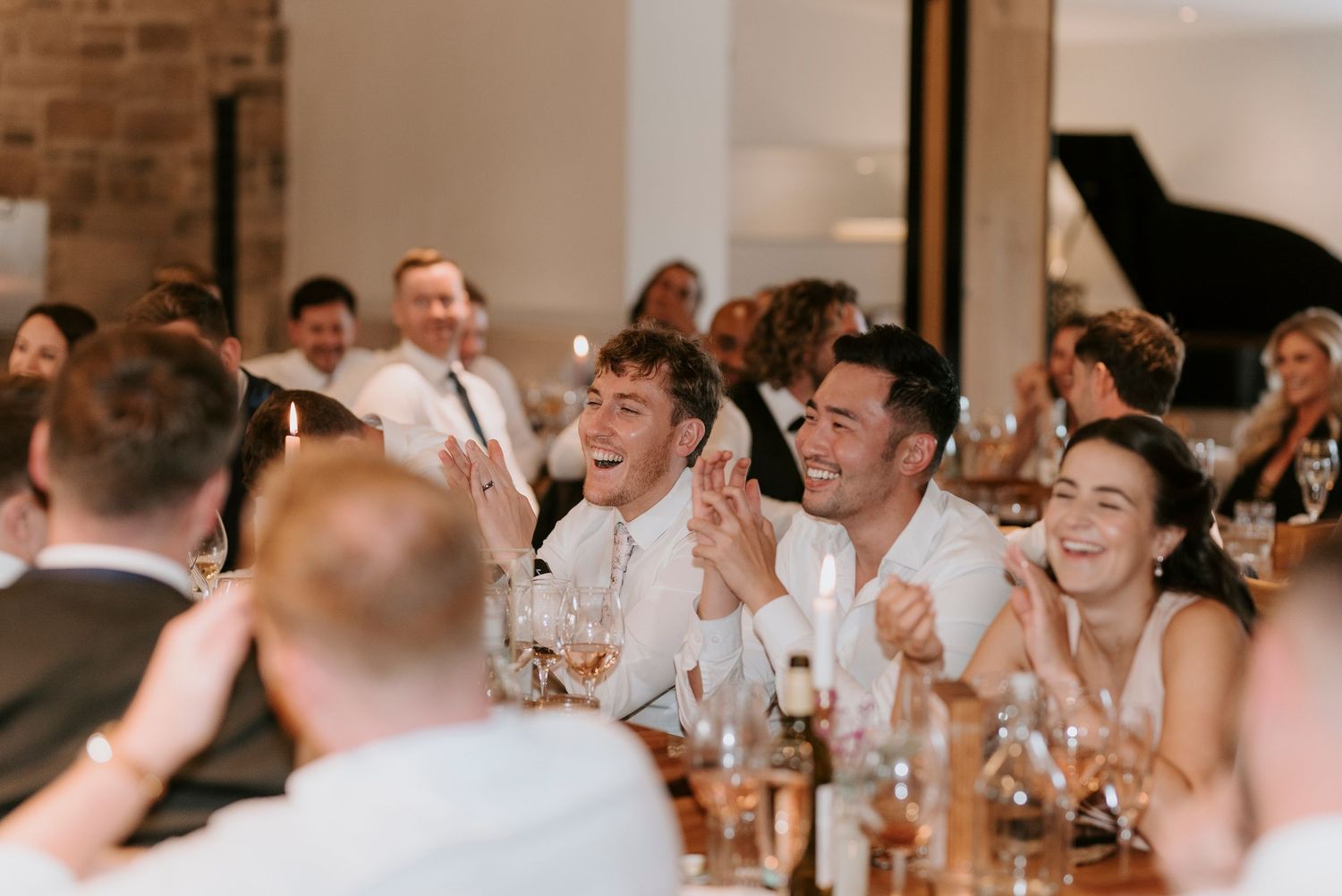 Wedding guests laugh and celebrate together during a reception dinner at a rustic venue with brick walls.
