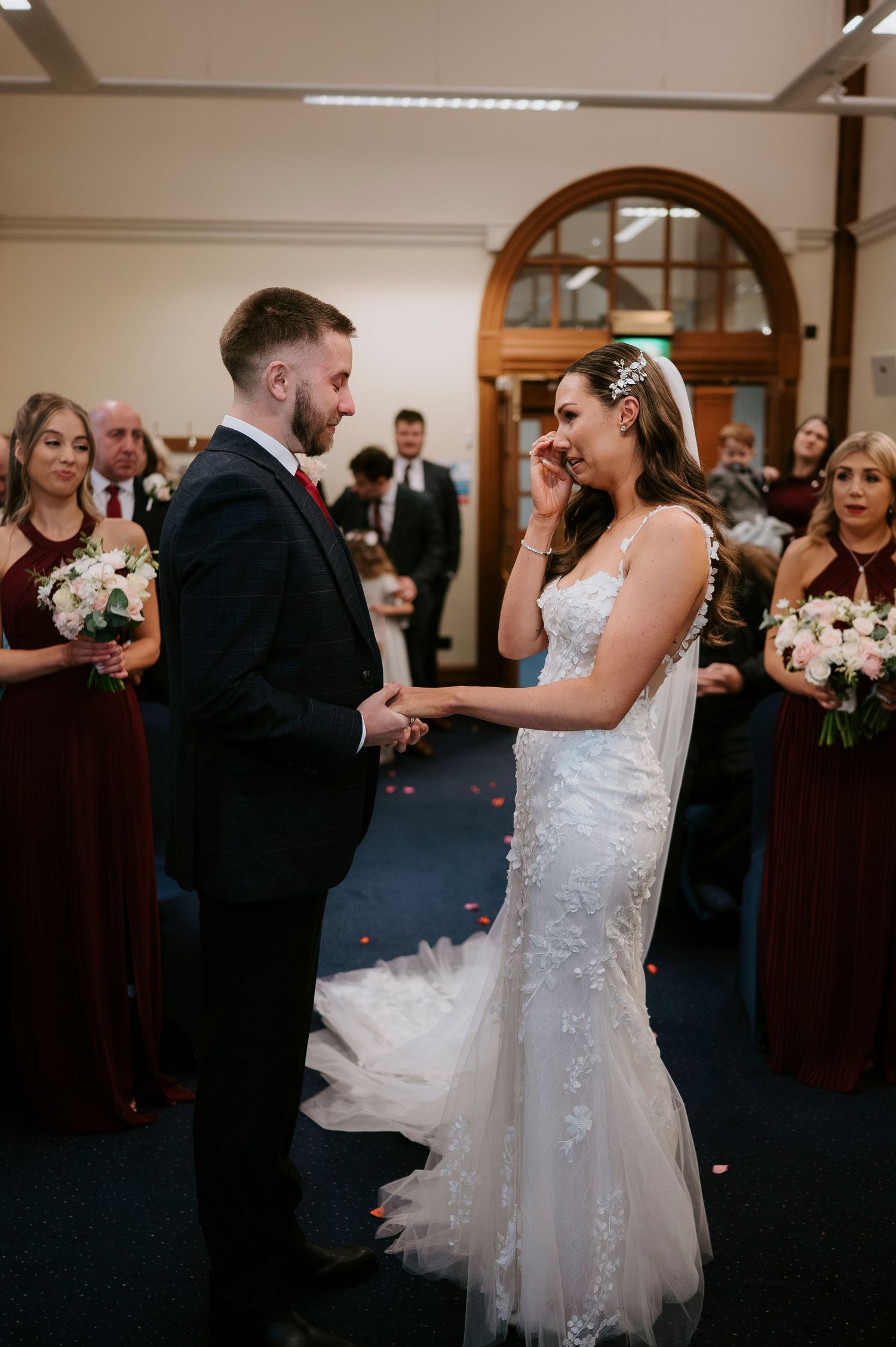 Bride wiping tears while holding hands with groom during emotional wedding ceremony, bridesmaids in burgundy dresses behind.