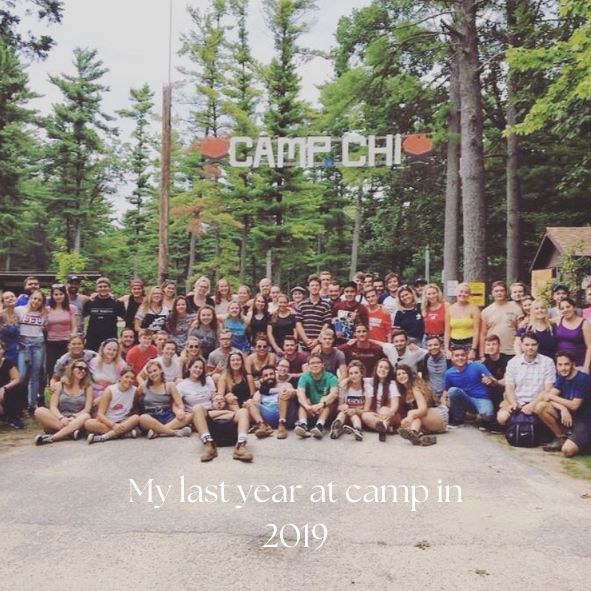 Large group photo taken at Camp Chi in 2019 with tall pine trees and wooden camp sign visible in background.
