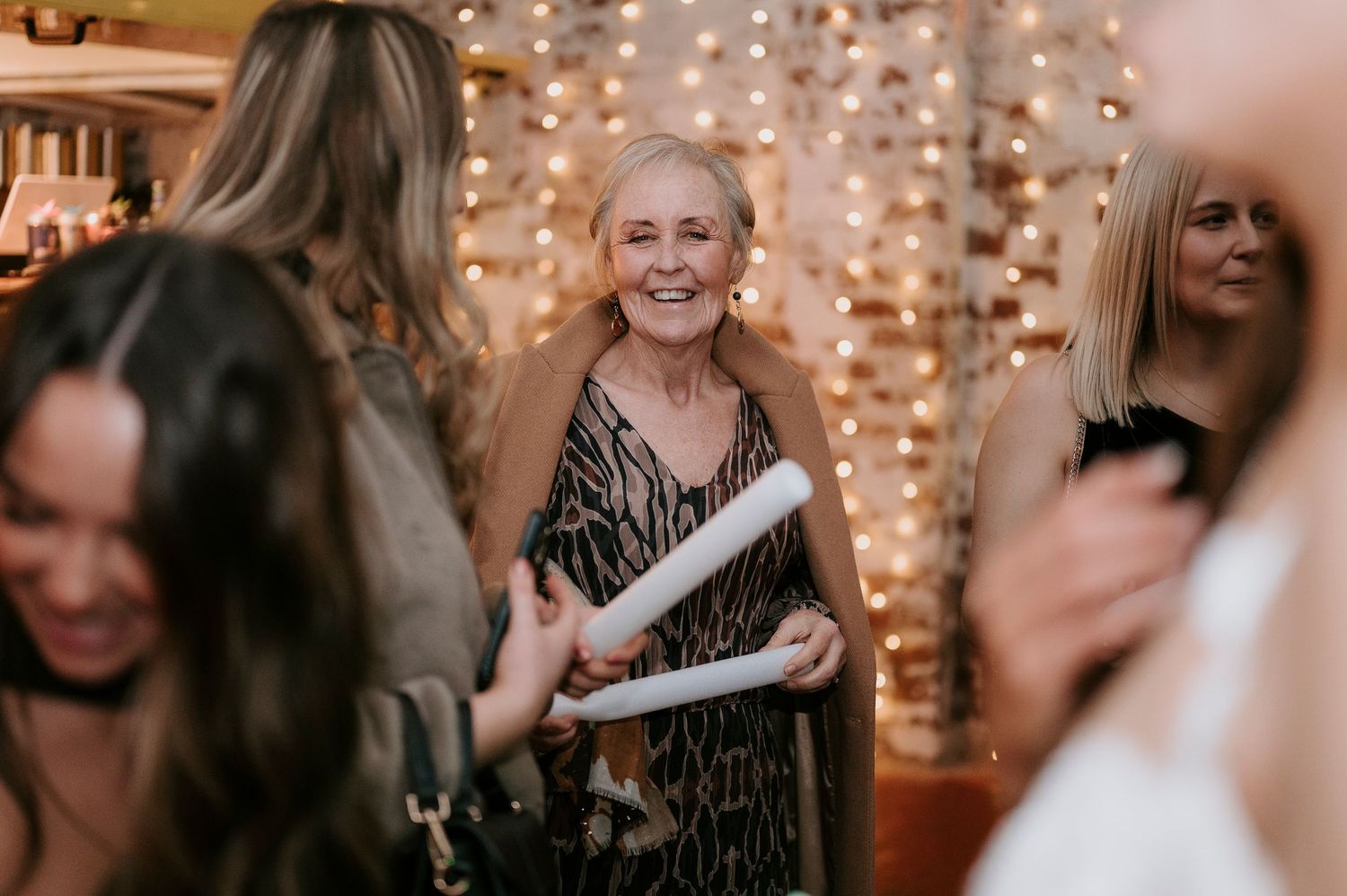Smiling older woman holding rolled paper surrounded by guests in front of fairy light backdrop.
