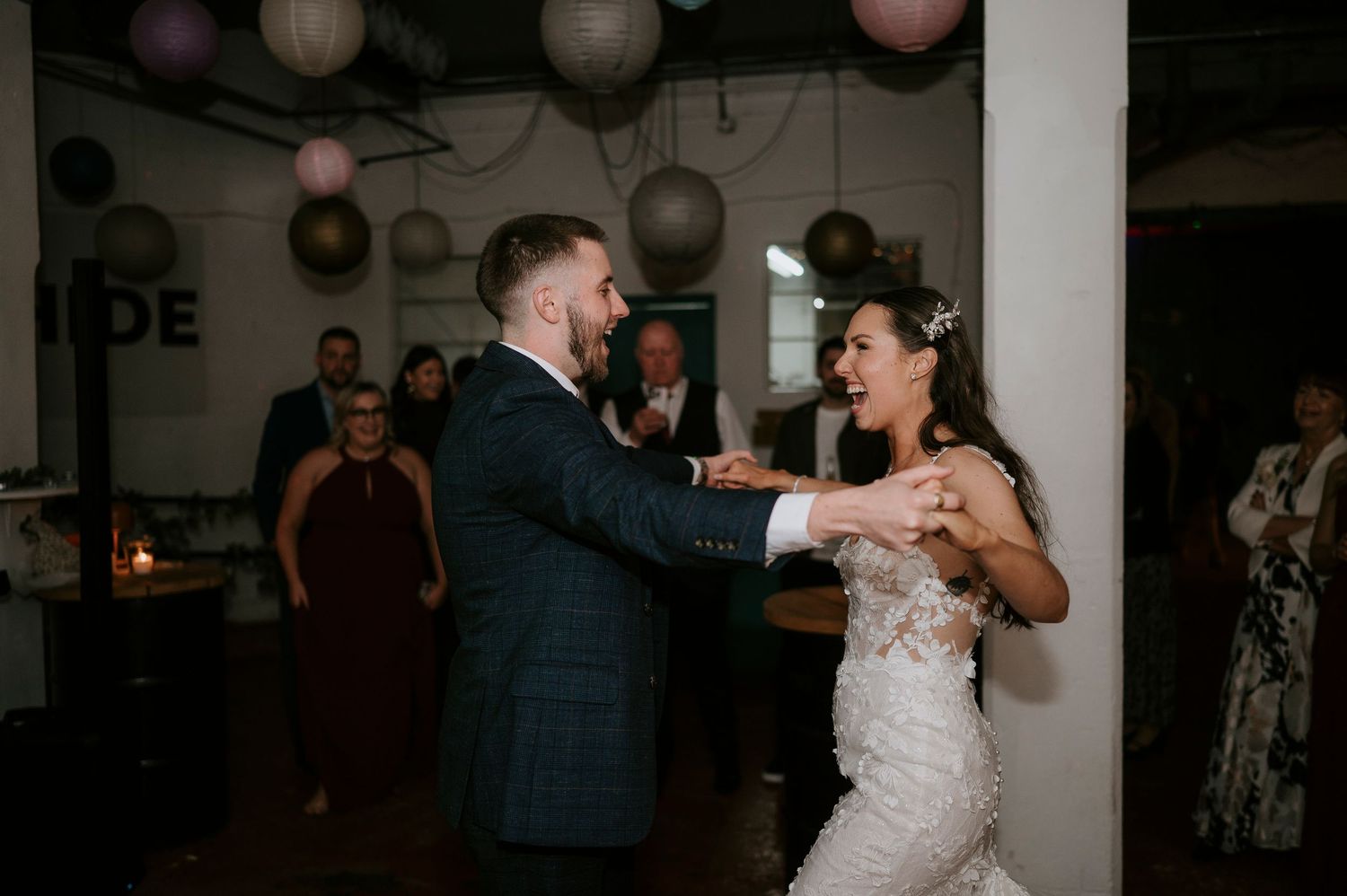 Bride and groom dance joyfully at wedding reception surrounded by guests and paper lanterns.