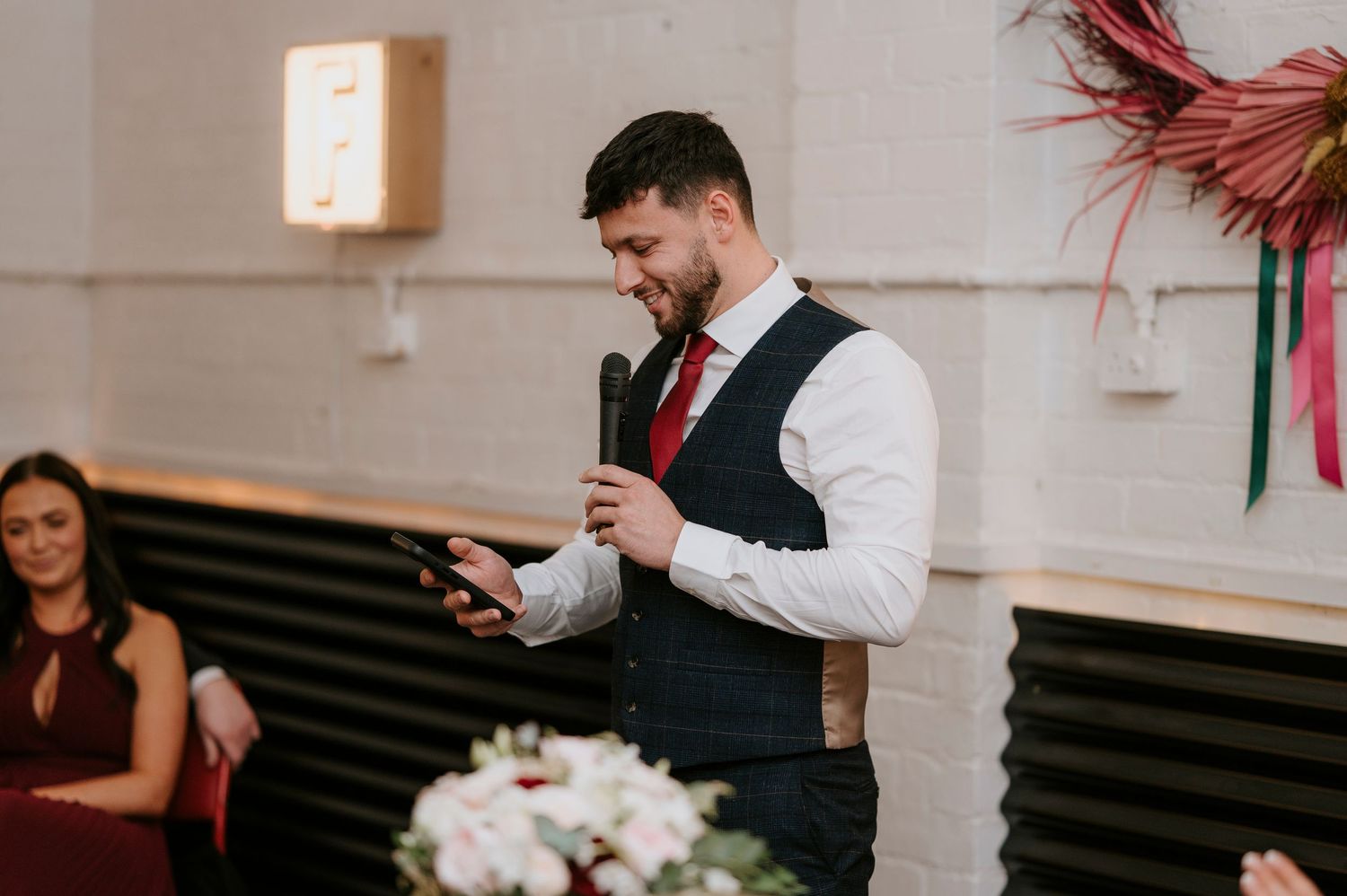 Groomsman in navy vest and red tie delivering speech with microphone at wedding reception.