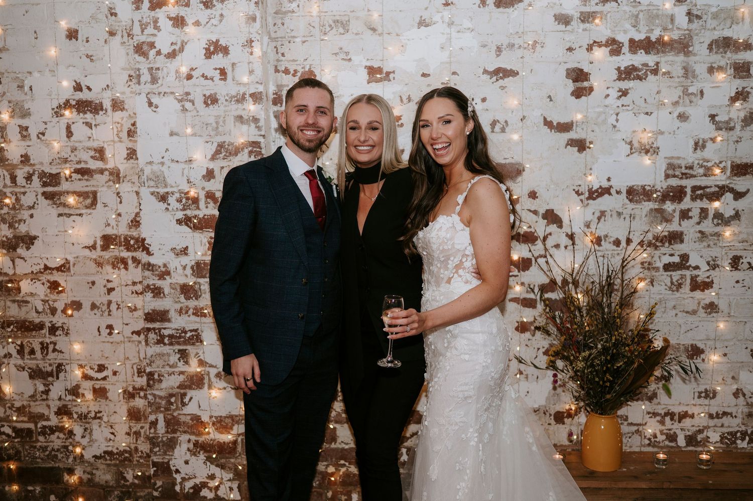 Bride and groom pose with wedding celebrant in front of fairy light brick wall at wedding reception.