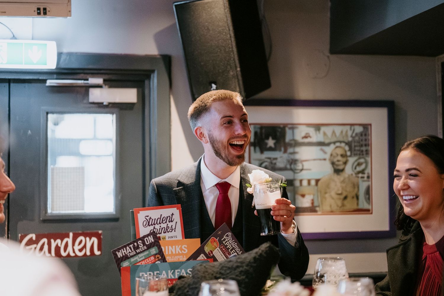 A smiling man in a suit raises a cocktail glass at a lively bar, surrounded by laughing guests.
