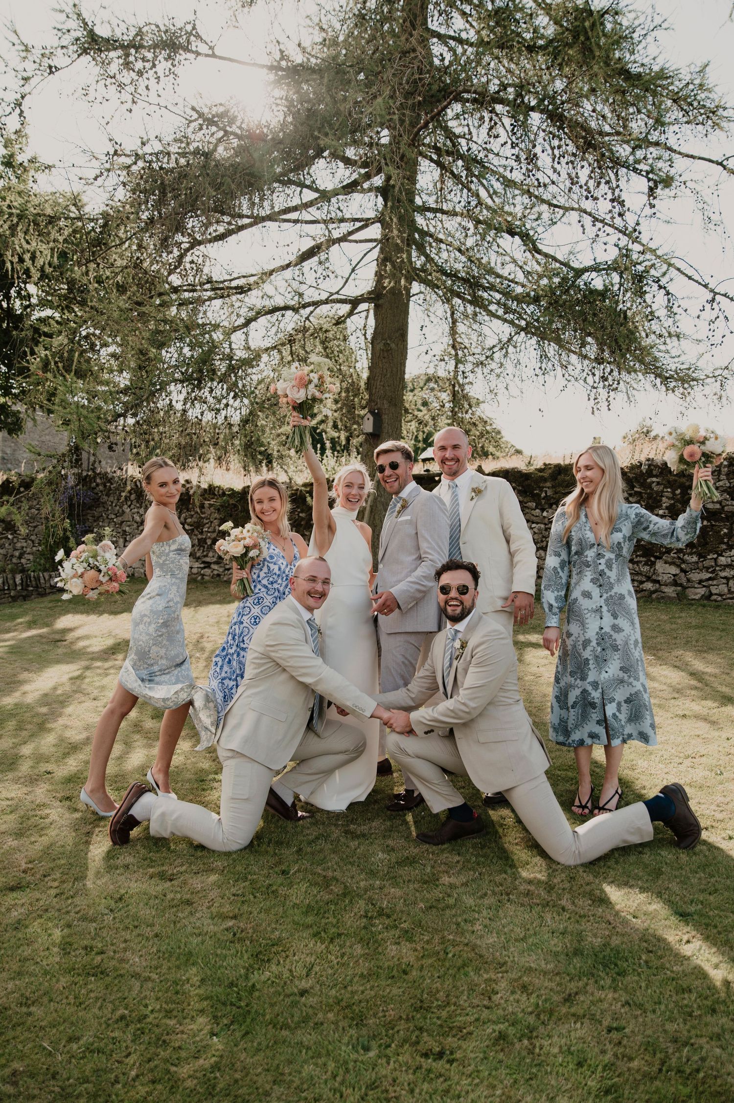 Fun wedding party photo with coordinated poses on a sunny lawn with trees in background.