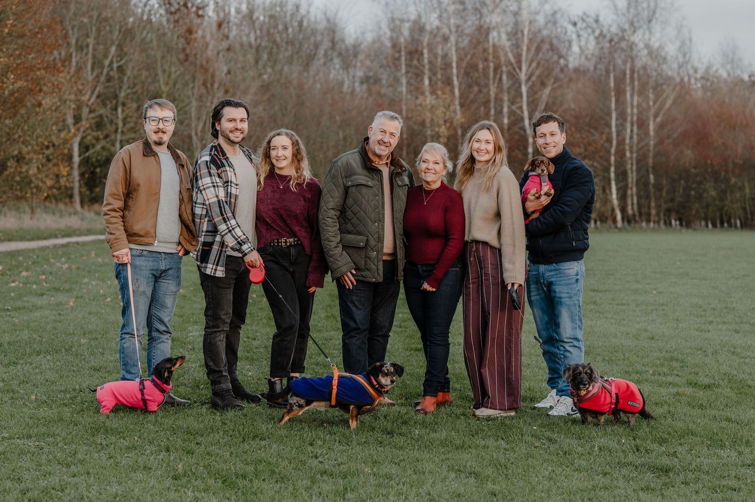 Group of people standing outdoors in casual fall attire with red duffel bags on grassy field during autumn.