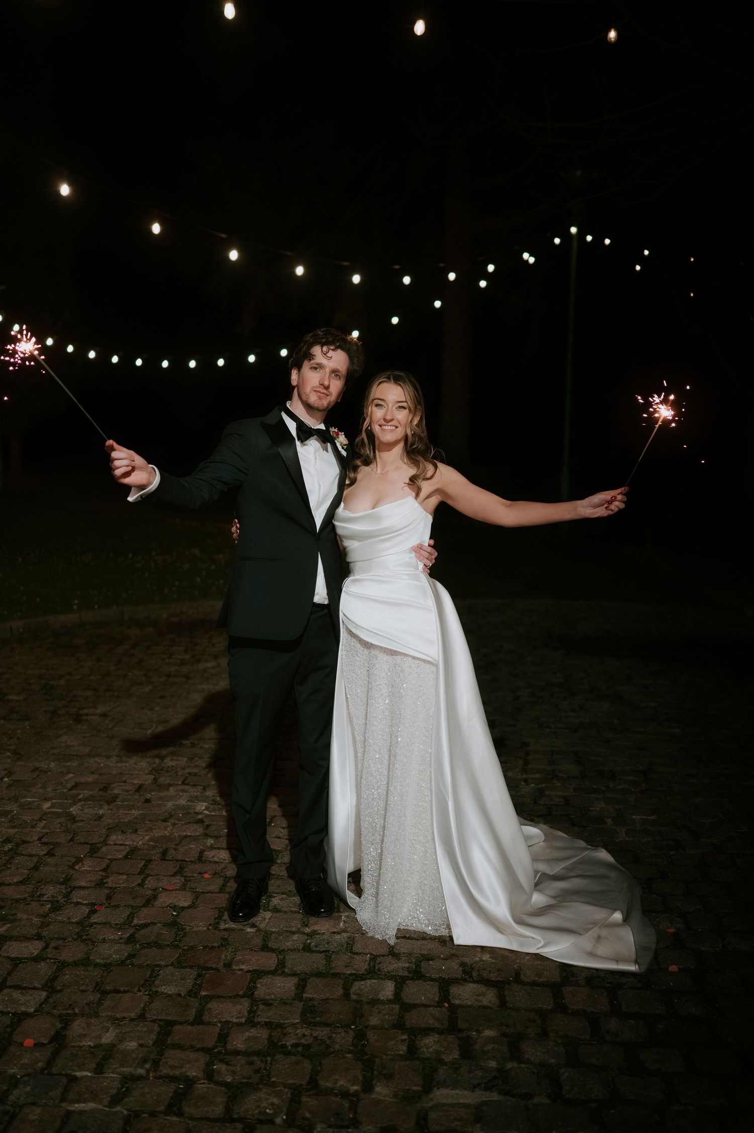 A couple holds sparklers while celebrating their wedding outdoors at night under string lights on a cobblestone path.