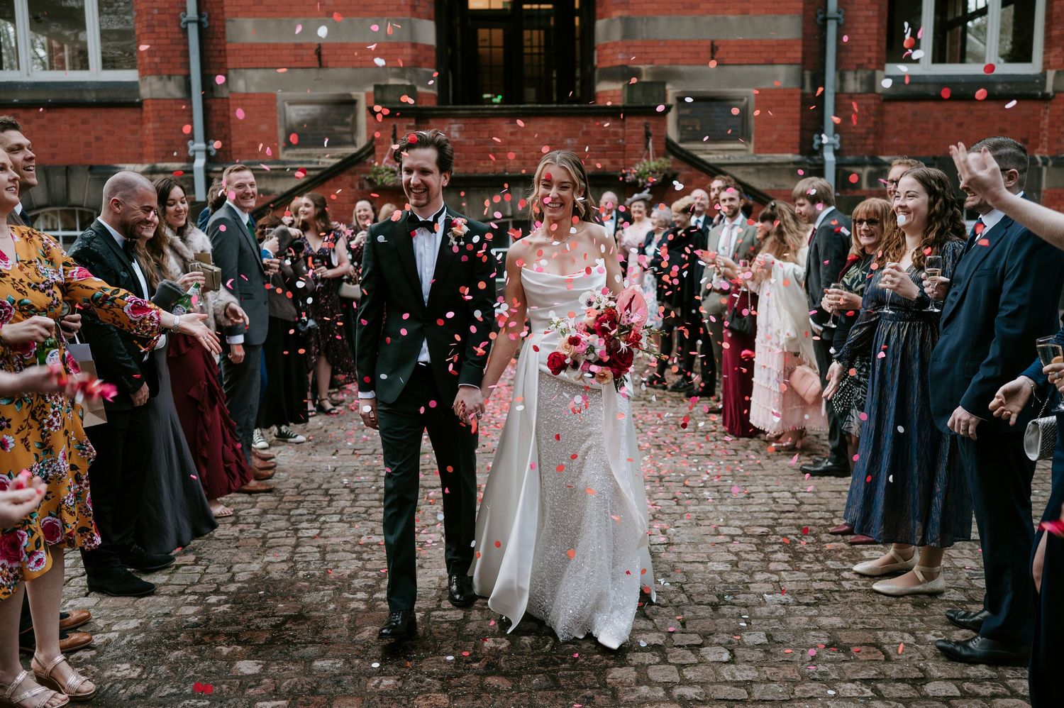 Wedding party stands on cobblestone path in front of historic red brick building during confetti toss.