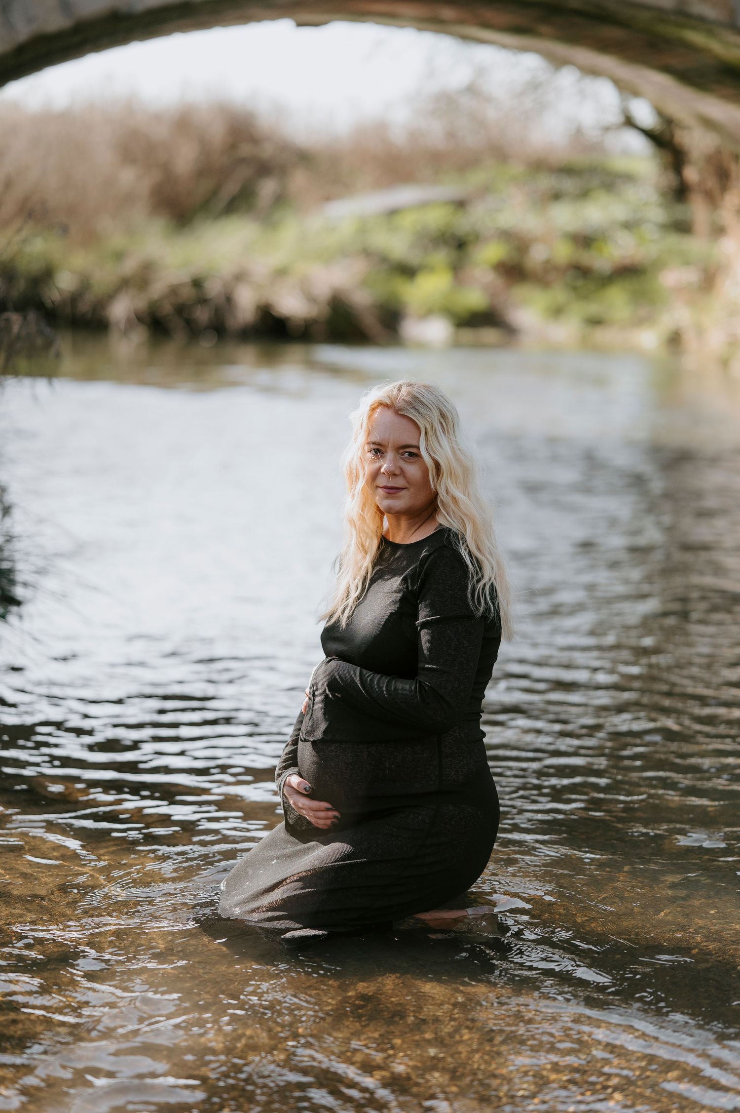 Artistic maternity photos taken in a river with the subject wearing a black dress and surrounded by water.