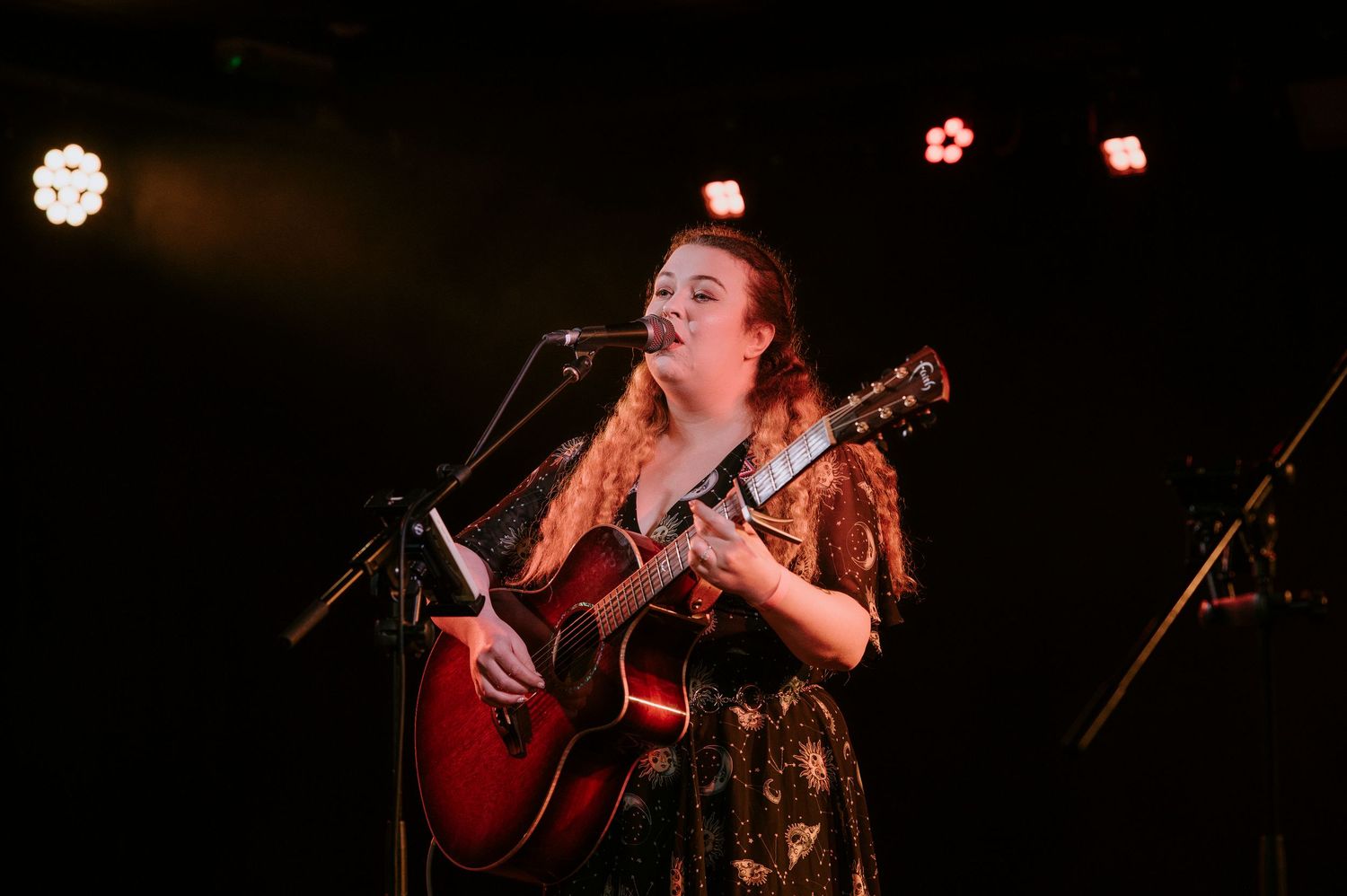 A performer sings and plays acoustic guitar on a dimly lit stage during a live concert performance.
