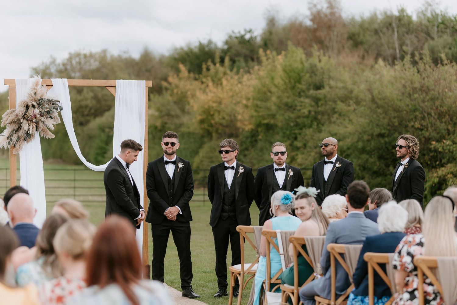 Wedding party stands in a line outdoors during a ceremony with guests seated on white chairs in a garden setting.