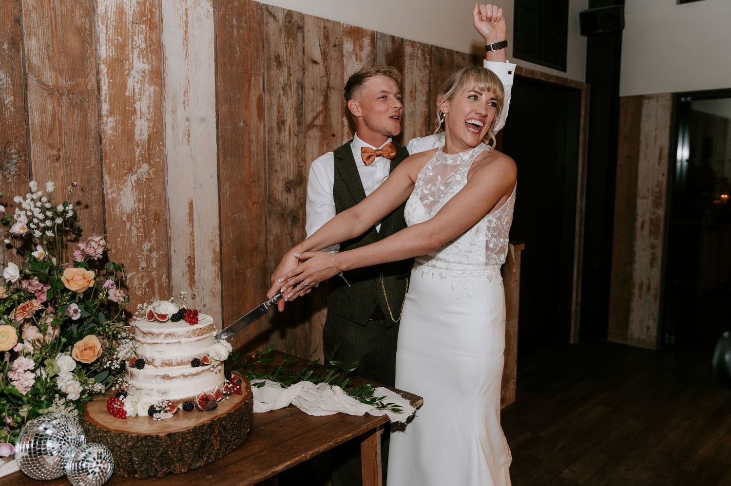 A newlywed couple cuts their elegant white wedding cake at a rustic wooden venue decorated with floral arrangements.