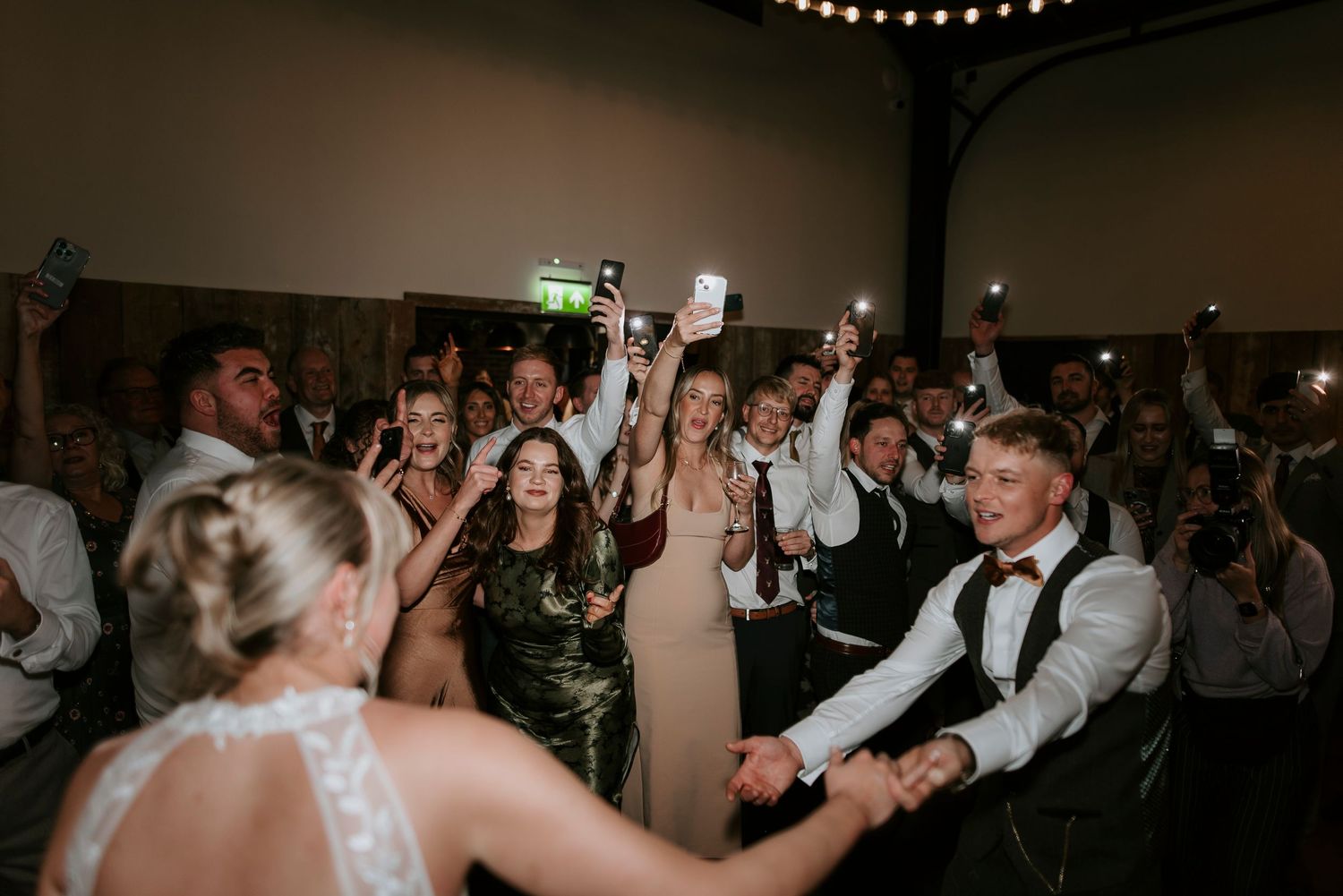 Wedding reception guests dancing and celebrating on a crowded dance floor under indoor lighting.