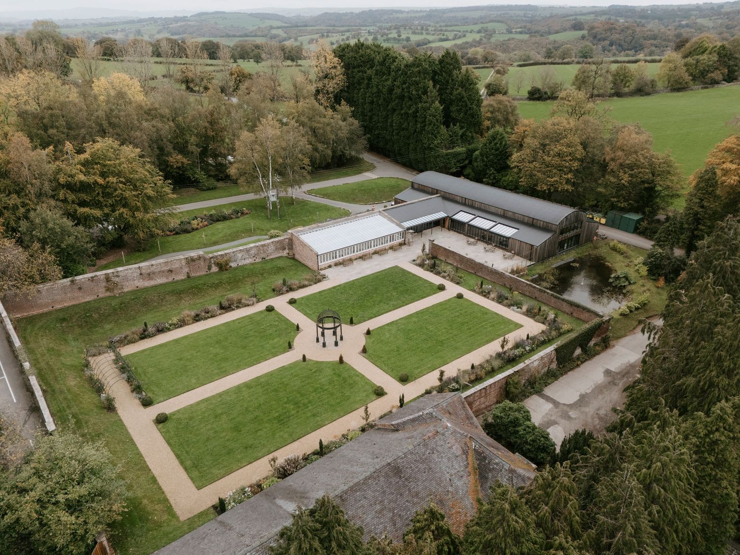 Aerial view of a formal garden with symmetrical paths and greenhouses surrounded by rolling countryside landscape.