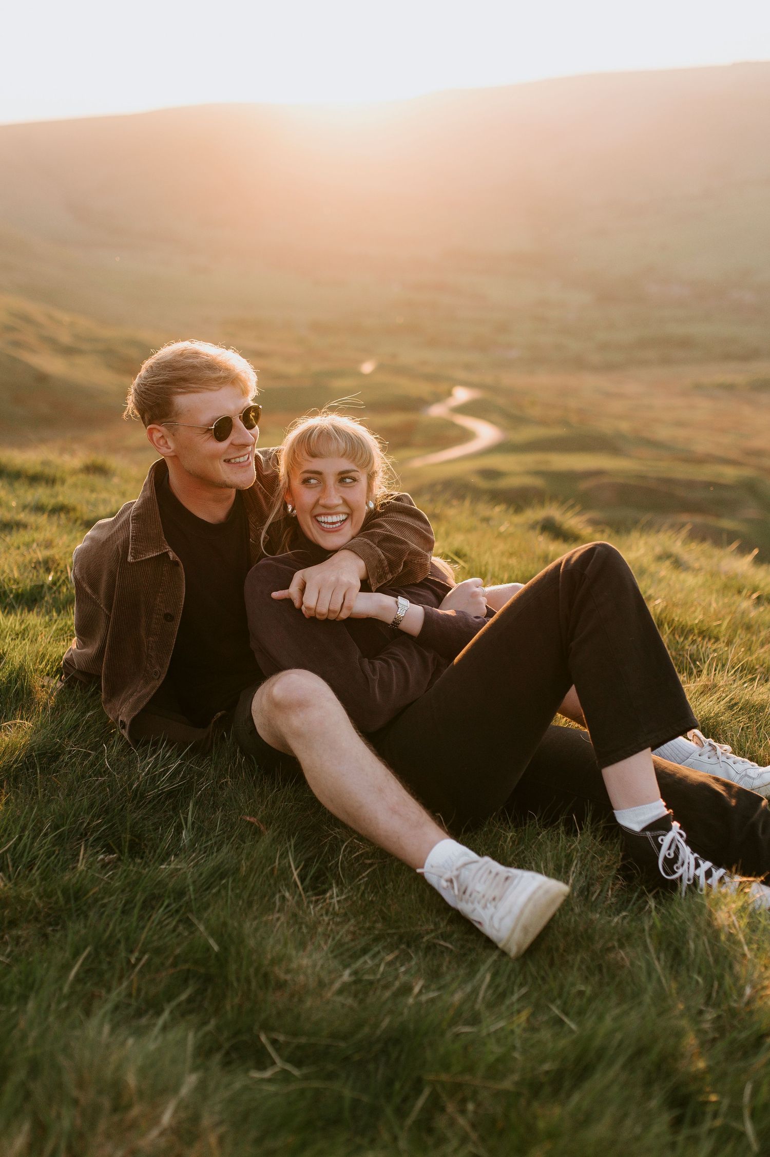 A couple lies together in golden sunlit grass on a hillside during sunset creating a romantic outdoor moment.