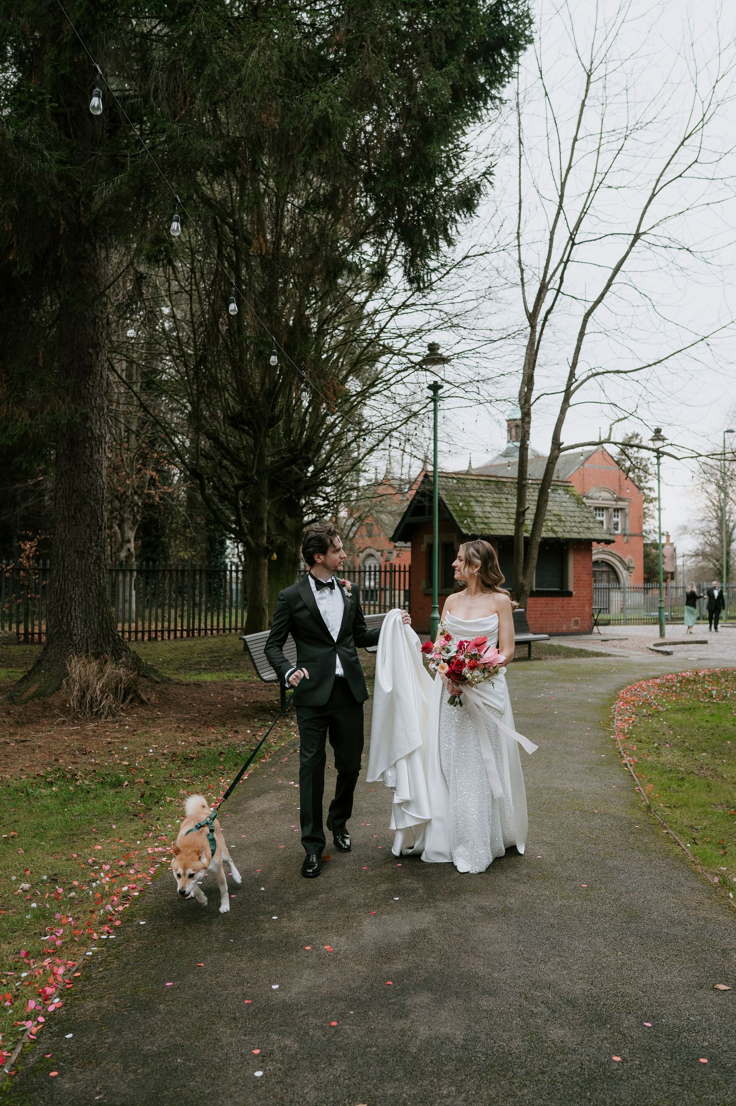 A wedding party walks along a tree-lined path on a cloudy day with scattered autumn leaves on the ground.