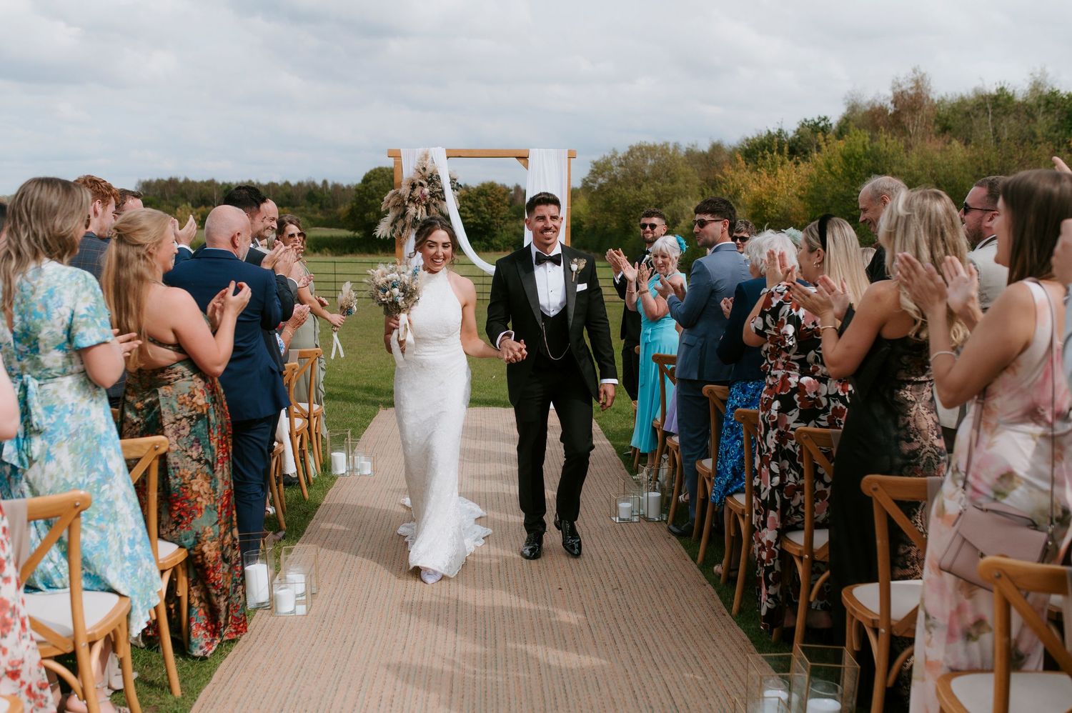 Newlyweds walk down outdoor ceremony aisle while wedding guests stand and celebrate their union.