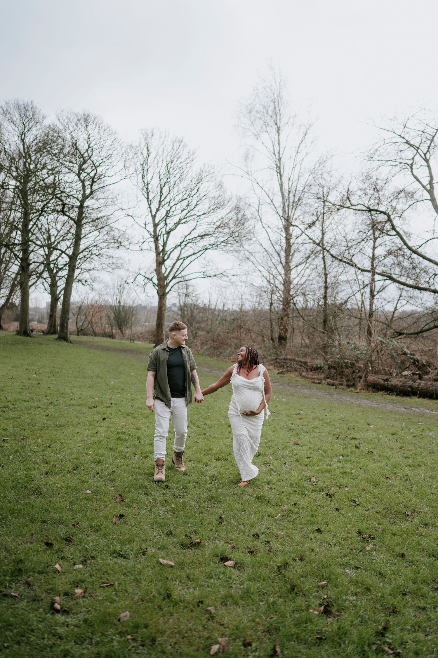 A couple walks hand in hand through a serene green meadow surrounded by bare winter trees on an overcast day.