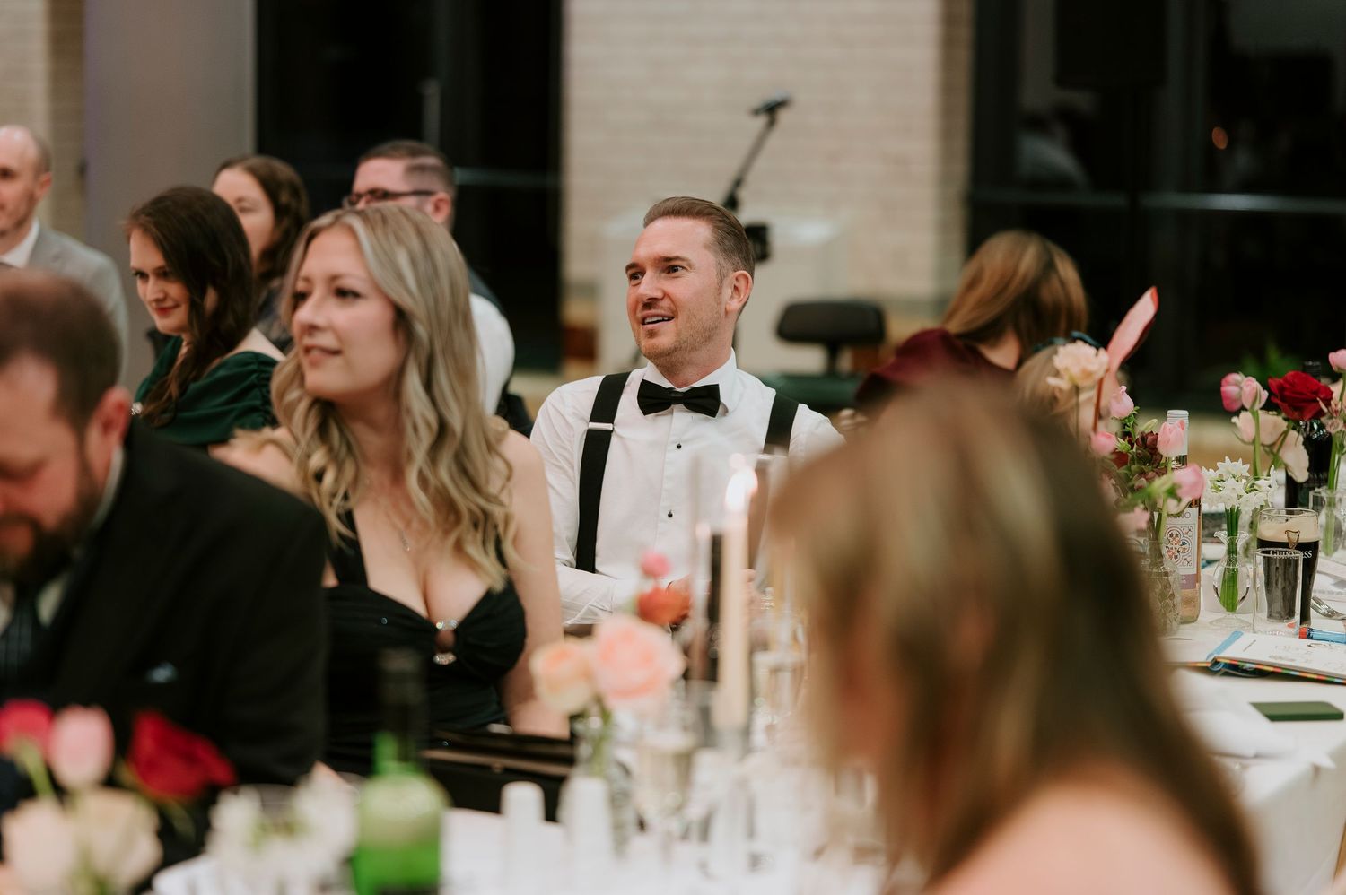 Wedding guests laugh and smile during dinner reception at elegant indoor venue with brick walls.