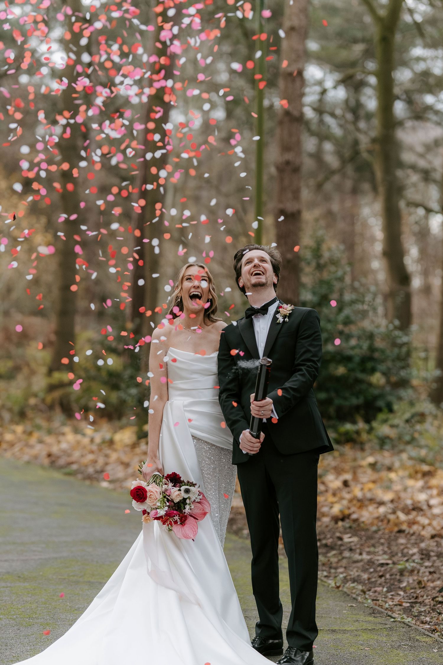 A couple celebrates their wedding day outdoors with pink flower petals falling around them in a wooded setting.
