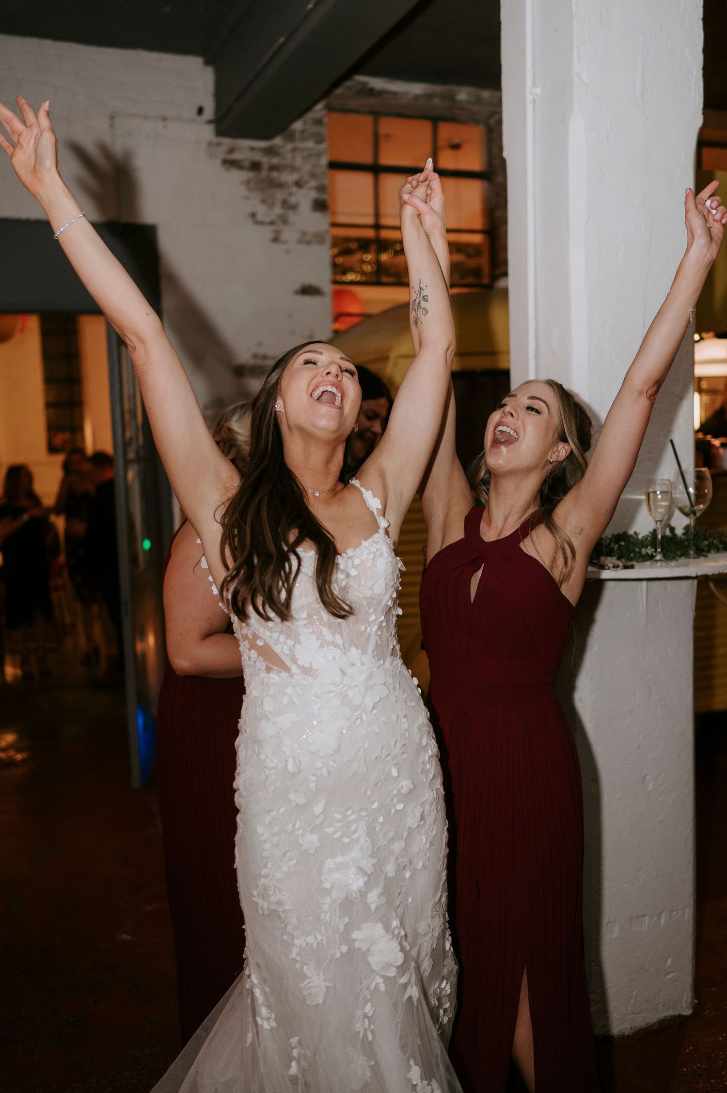 Bride and bridesmaid joyfully raise their arms on the wedding reception dance floor.