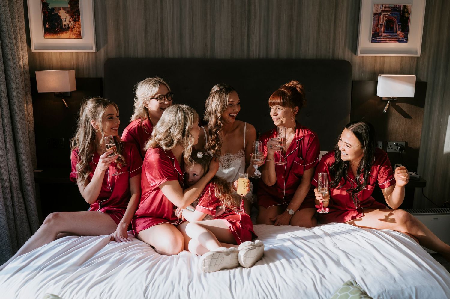 Six bridesmaids in red satin robes celebrate on a hotel bed, toasting with champagne glasses during bridal party preparations.
