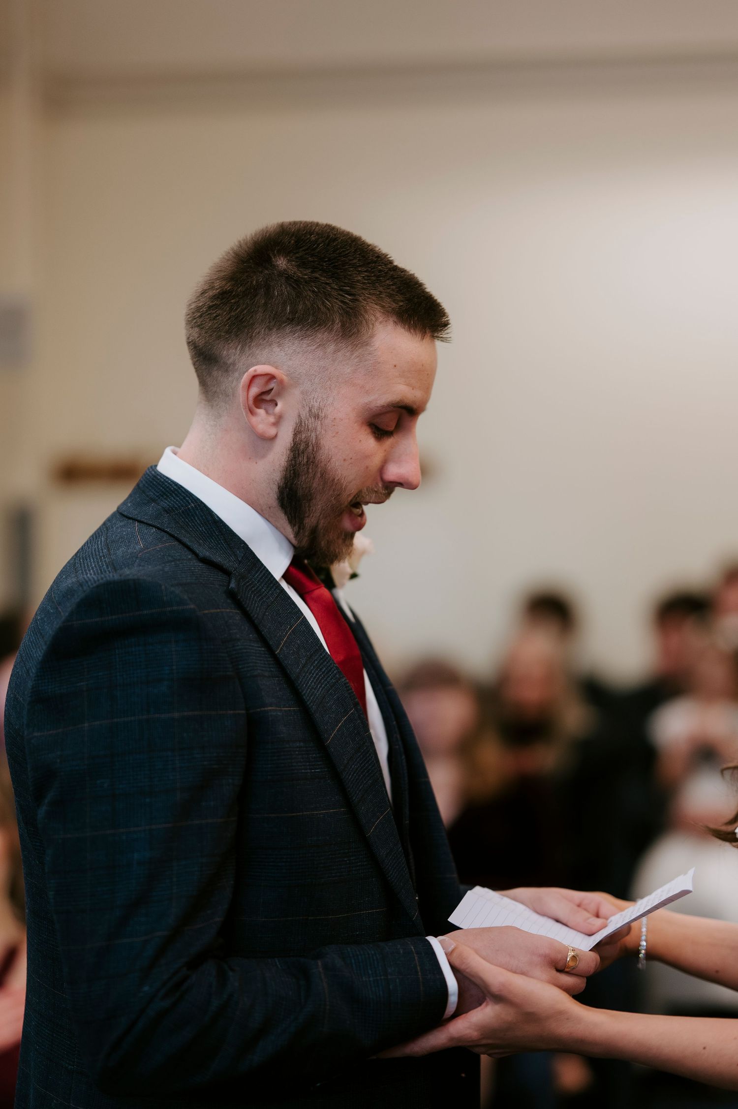 Groom in navy suit and red tie reading wedding vows during ceremony.