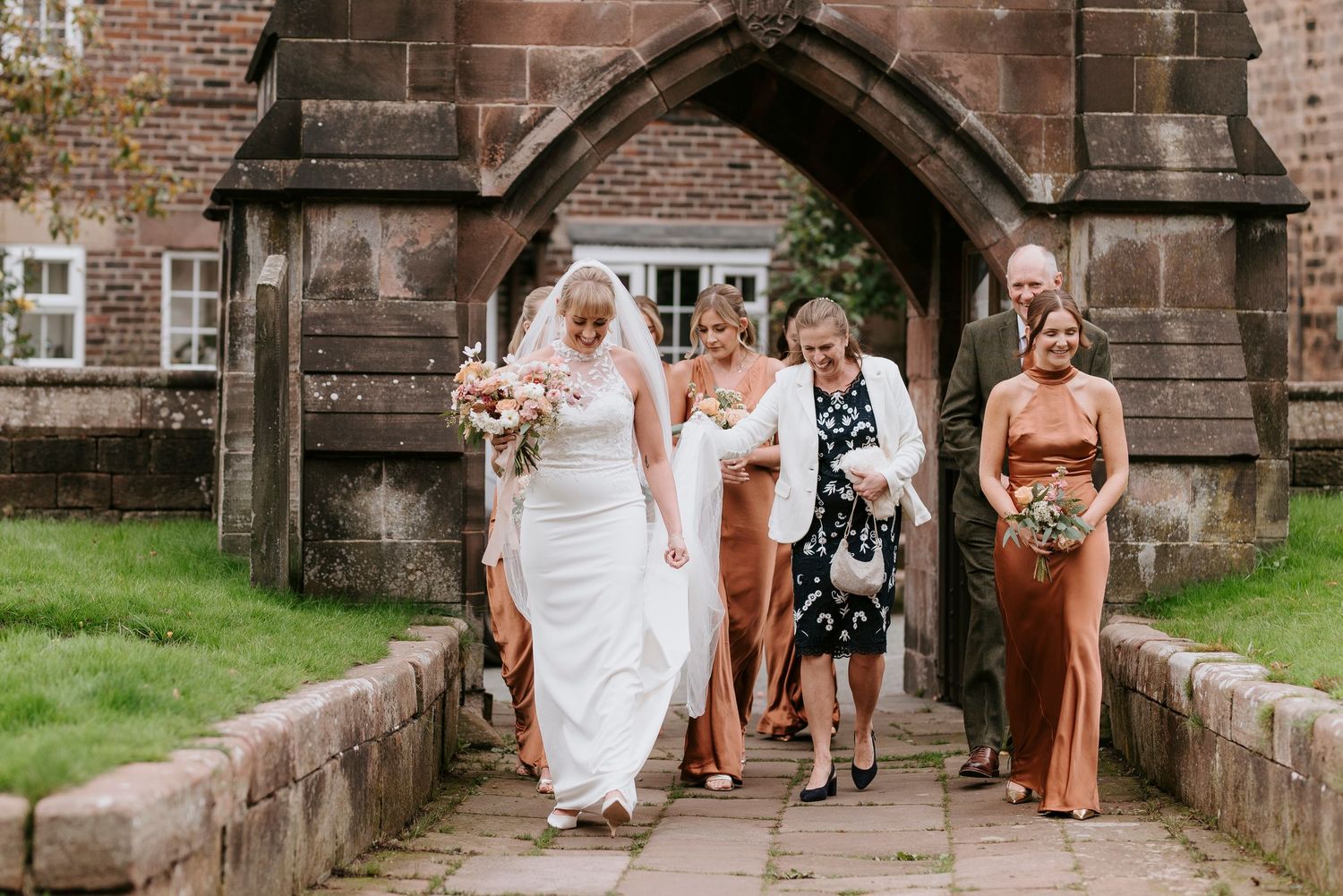 A wedding party walks along a stone church wall in a line with bridesmaids wearing brown dresses and carrying bouquets.