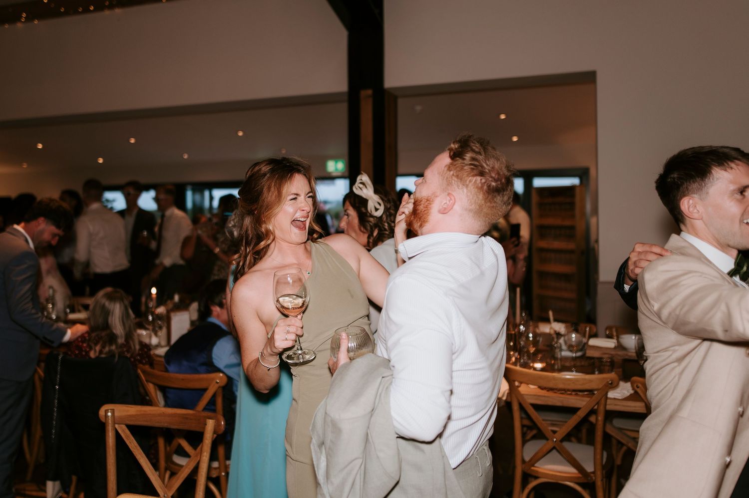 Wedding guests dance and laugh together during a lively reception celebration at an indoor venue.