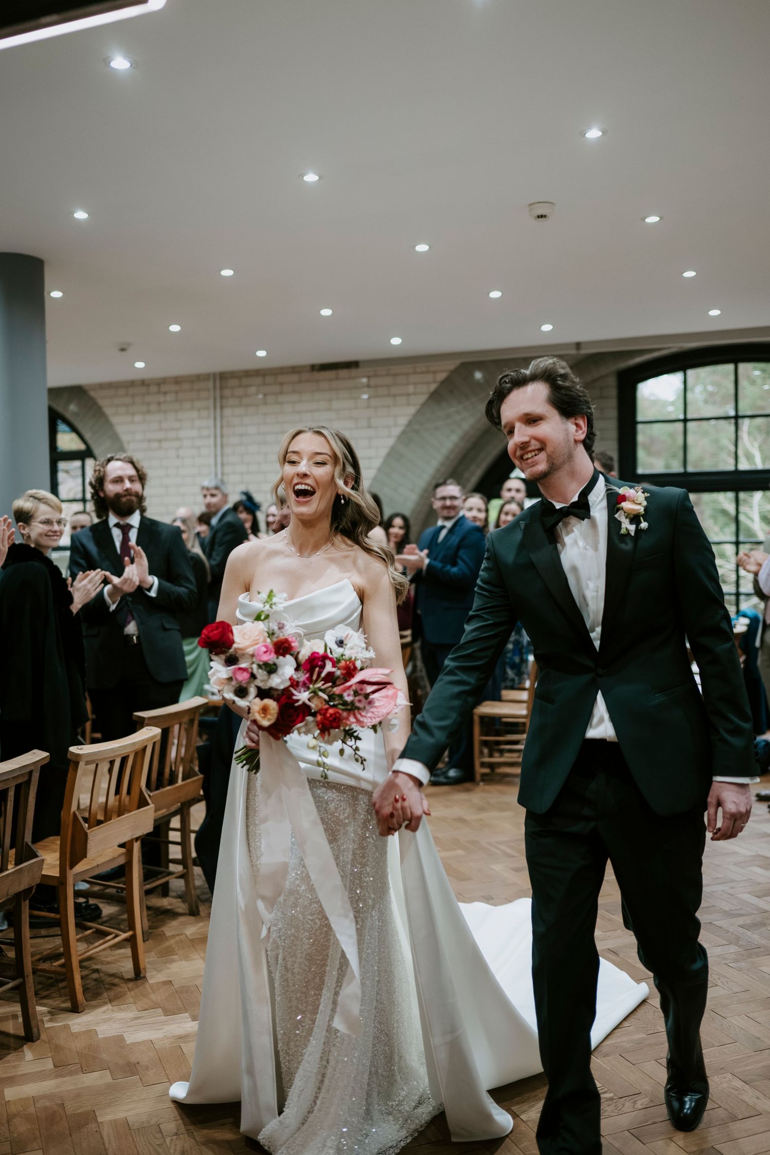 Wedding party exits church ceremony with joyful expressions and laughter while holding bouquets of red flowers.