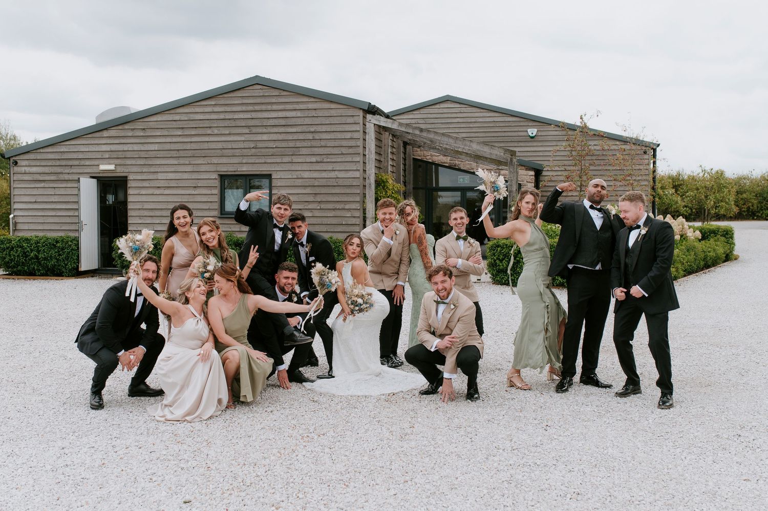 A wedding party poses together in a line outside a rustic building with gray siding.