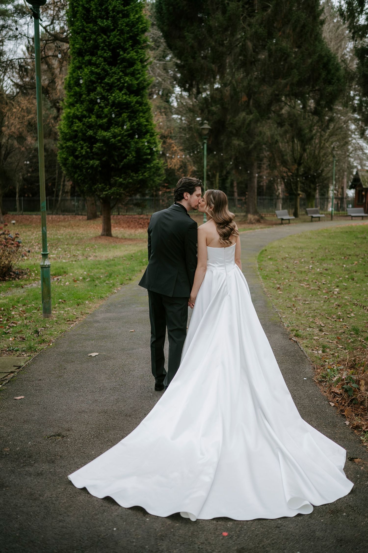 Newlyweds walk hand in hand through a park as the bride's dramatic white ballgown trails behind them.