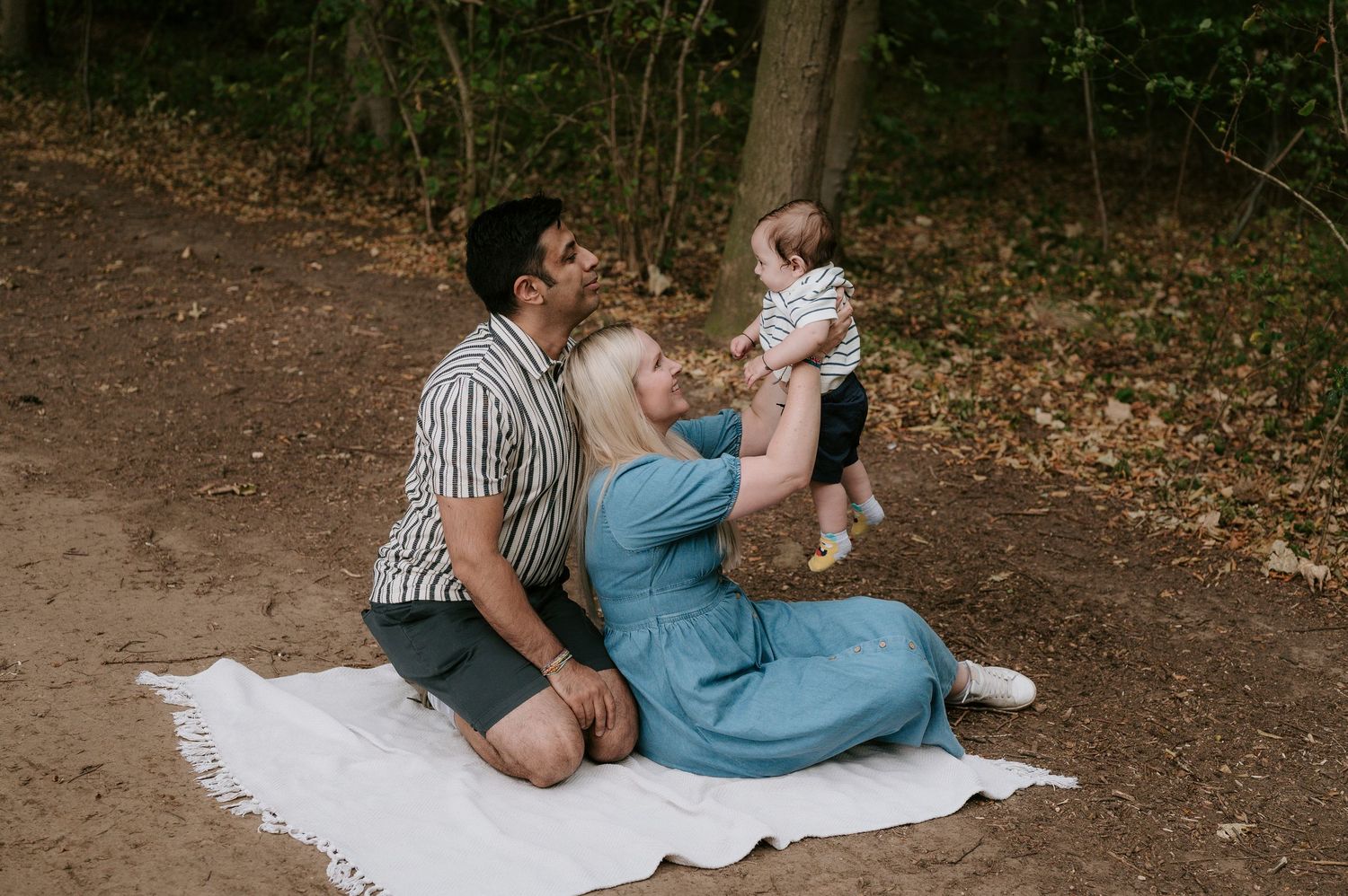Family sits together on white blanket during outdoor photoshoot in wooded area on dirt path.