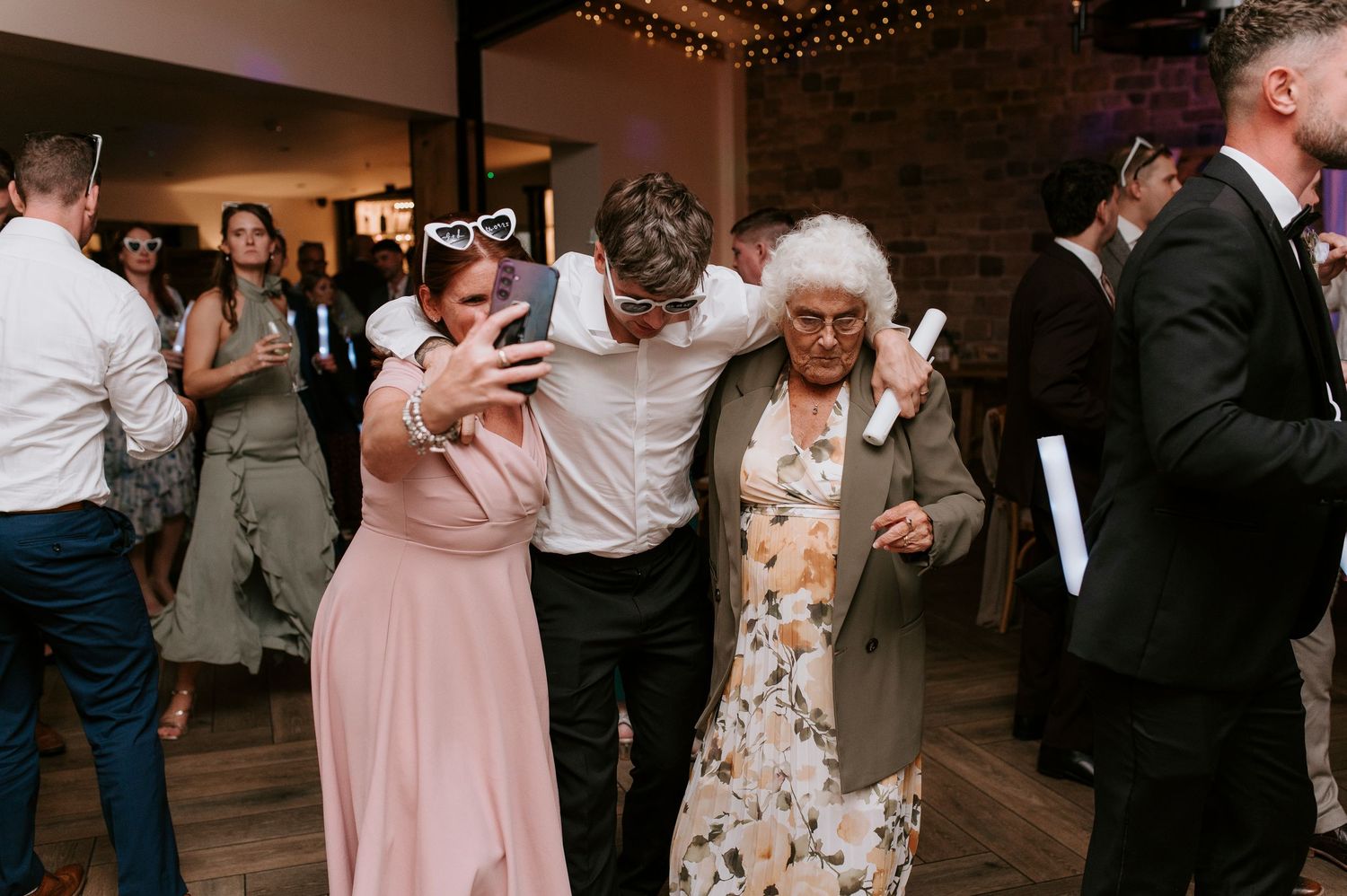 Wedding guests dance and celebrate on a rustic wooden dance floor under twinkling string lights at a reception.