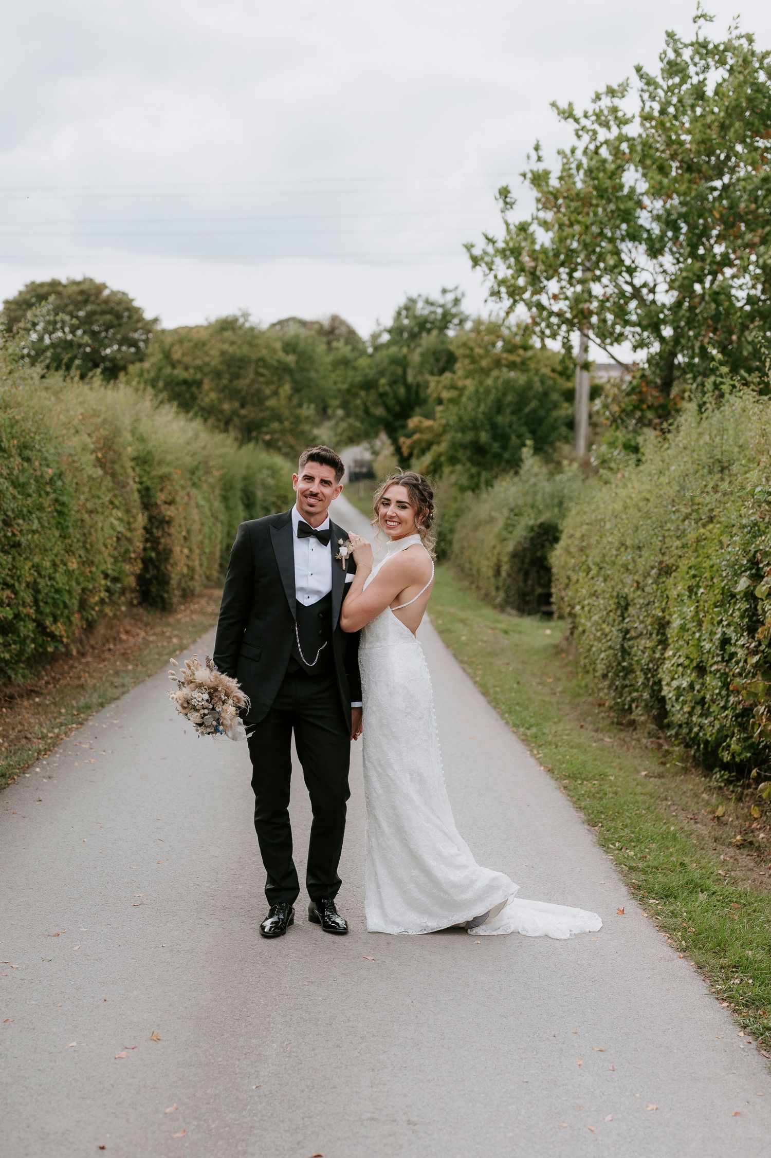 A couple in wedding attire walks and laughs together on a tree-lined path during their special day.