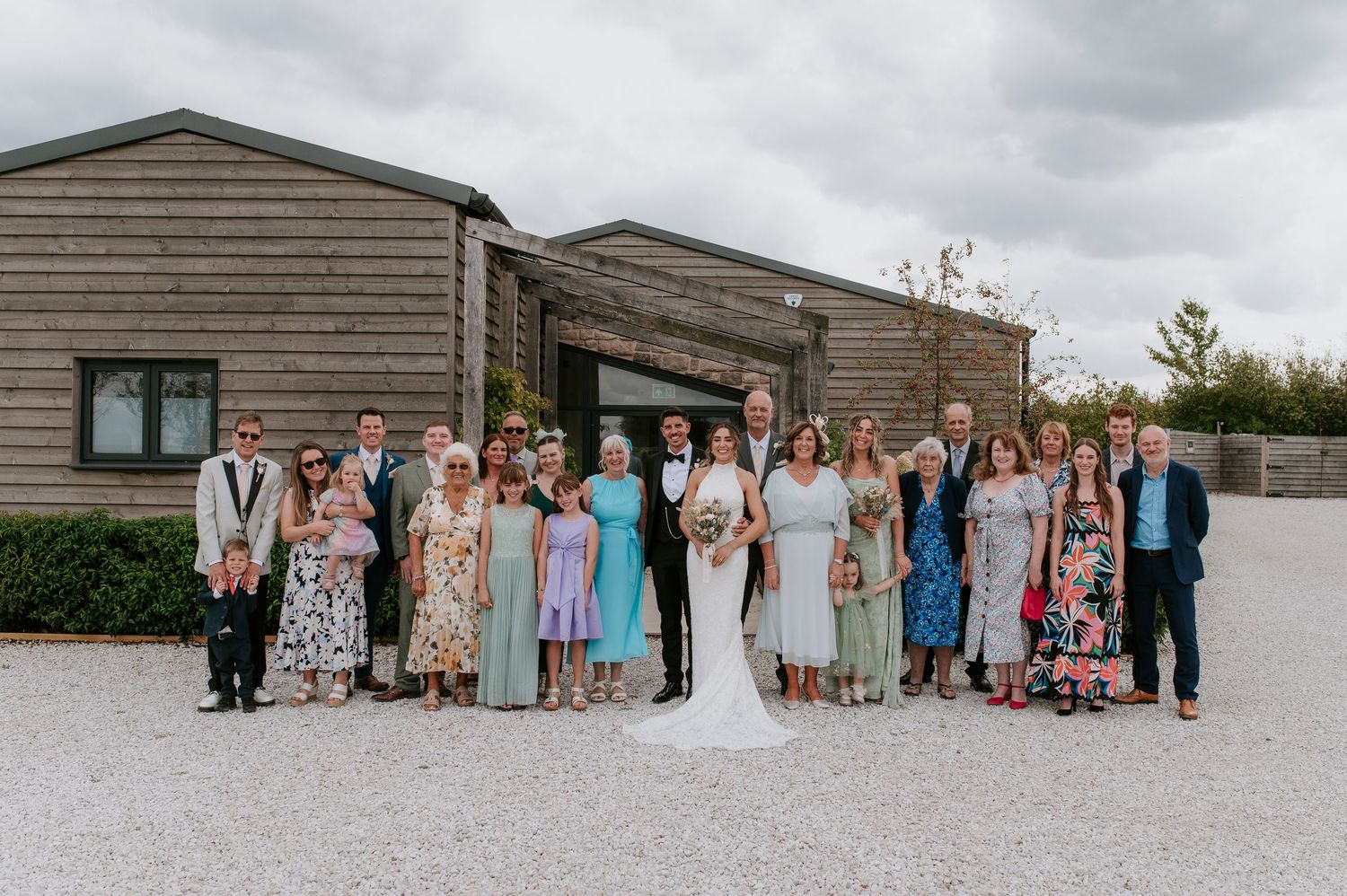 A large group of wedding guests and wedding party members pose together outside a rustic wooden building.