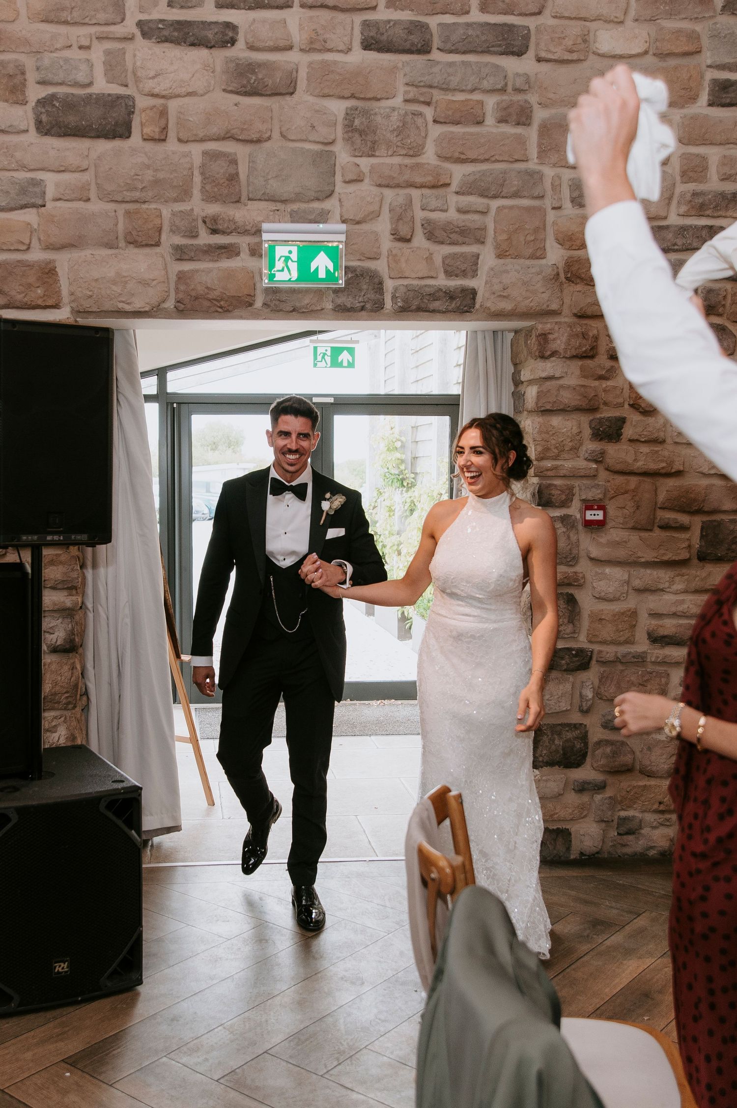 Wedding couple enters reception venue with joyful dance moves across a rustic brick-walled room with natural lighting.