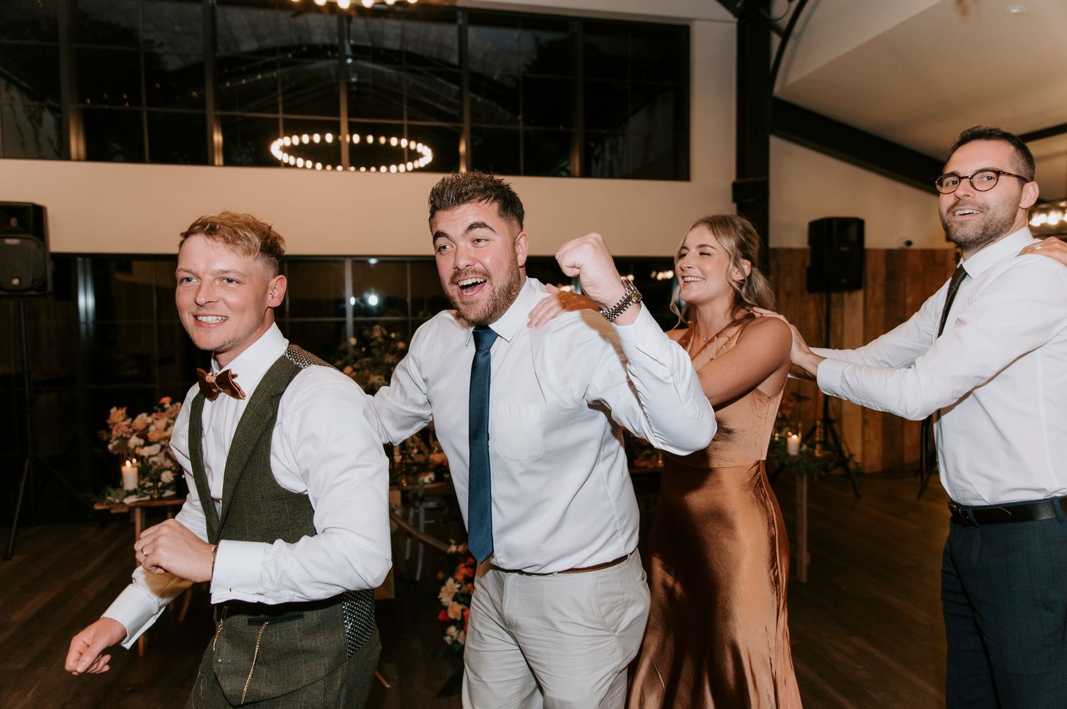 A group of wedding guests dance and celebrate together on a dimly lit dance floor at an evening reception.