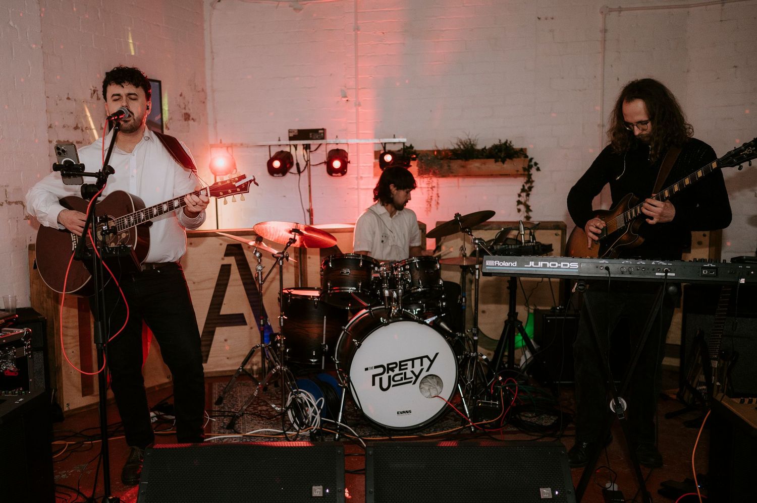 A three-piece band performs live, featuring two guitarists and a drummer on a small indoor stage with red ambient lighting.
