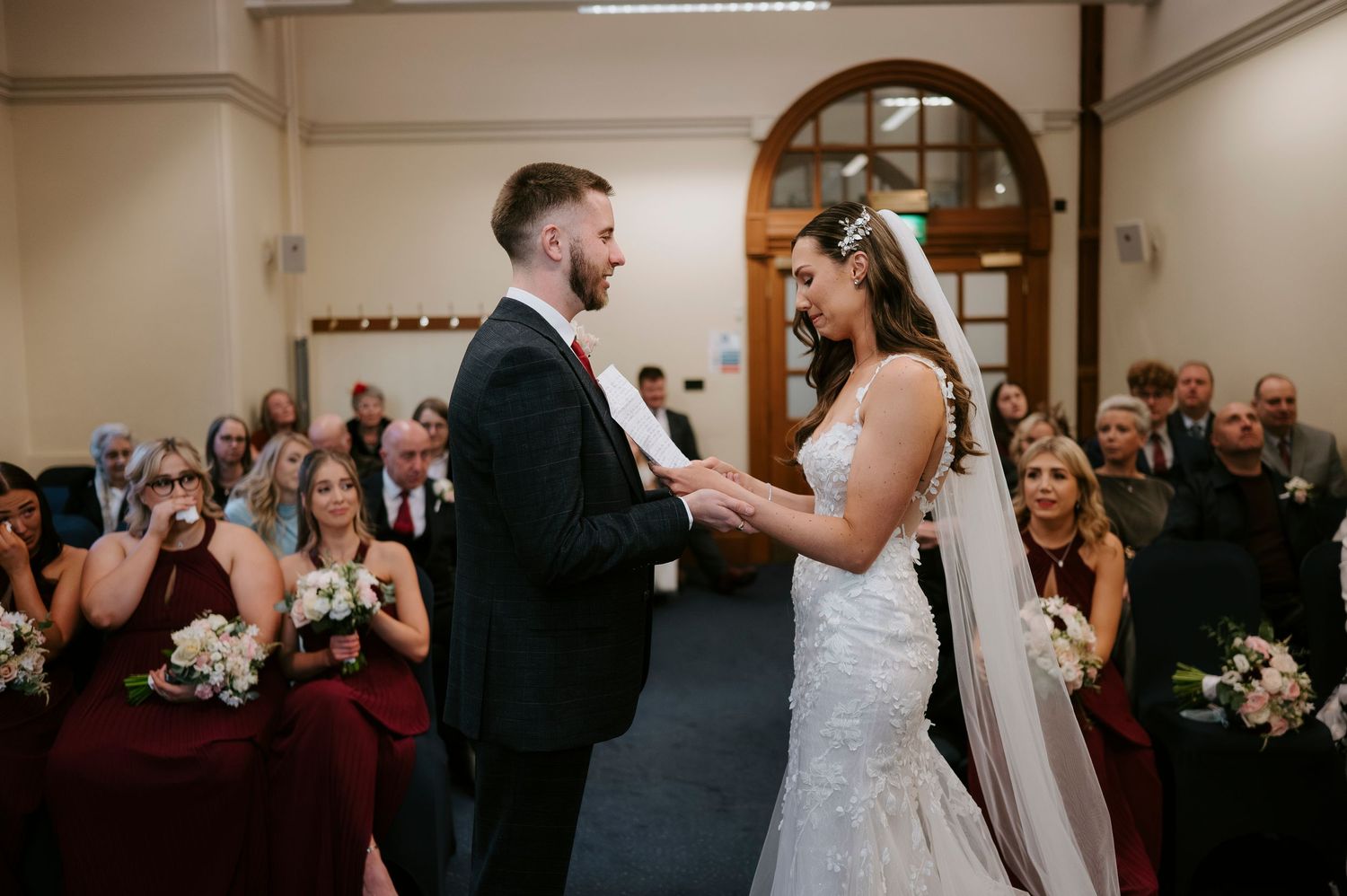Bride and groom exchange vows at wedding ceremony with bridesmaids in burgundy dresses and guests seated in background.