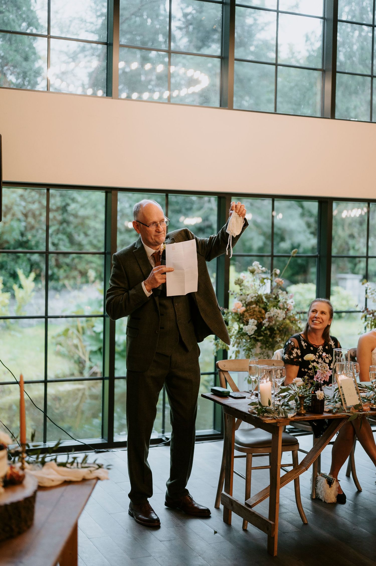 Emotional moment captured at a wedding reception in a modern glass-walled venue with natural lighting and elegant table settings.