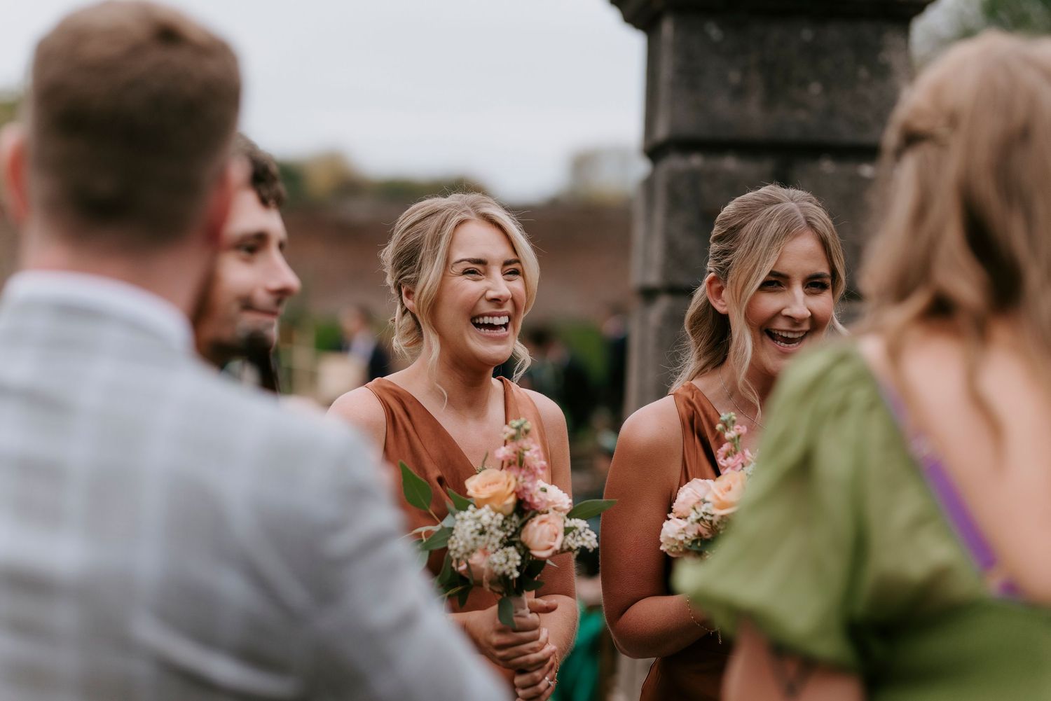 Wedding party members laugh together outdoors while holding bouquets during golden hour celebration.