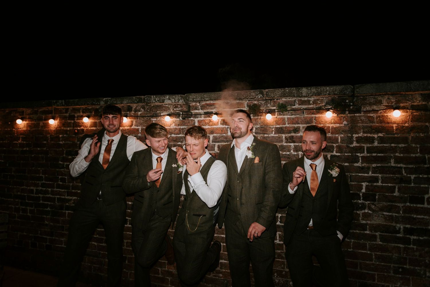 Wedding party members in formal attire pose together in front of a rustic brick wall with warm lighting.