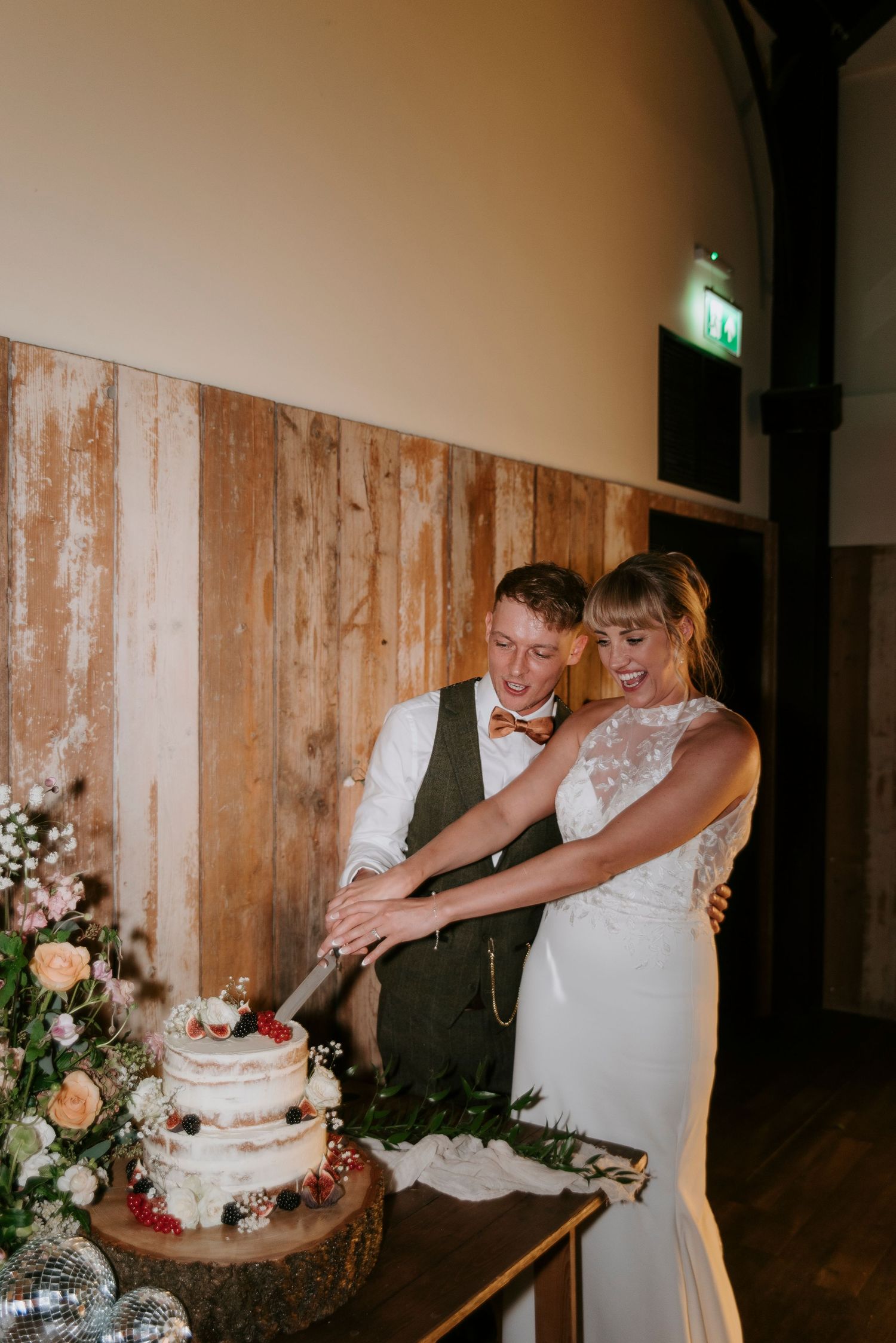 Couple cuts their rustic wedding cake together on wooden stand with floral decorations at reception.