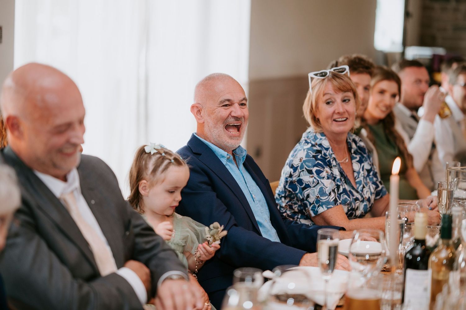 Wedding guests smile and laugh while seated at elegant reception tables with glowing candles and floral centerpieces.