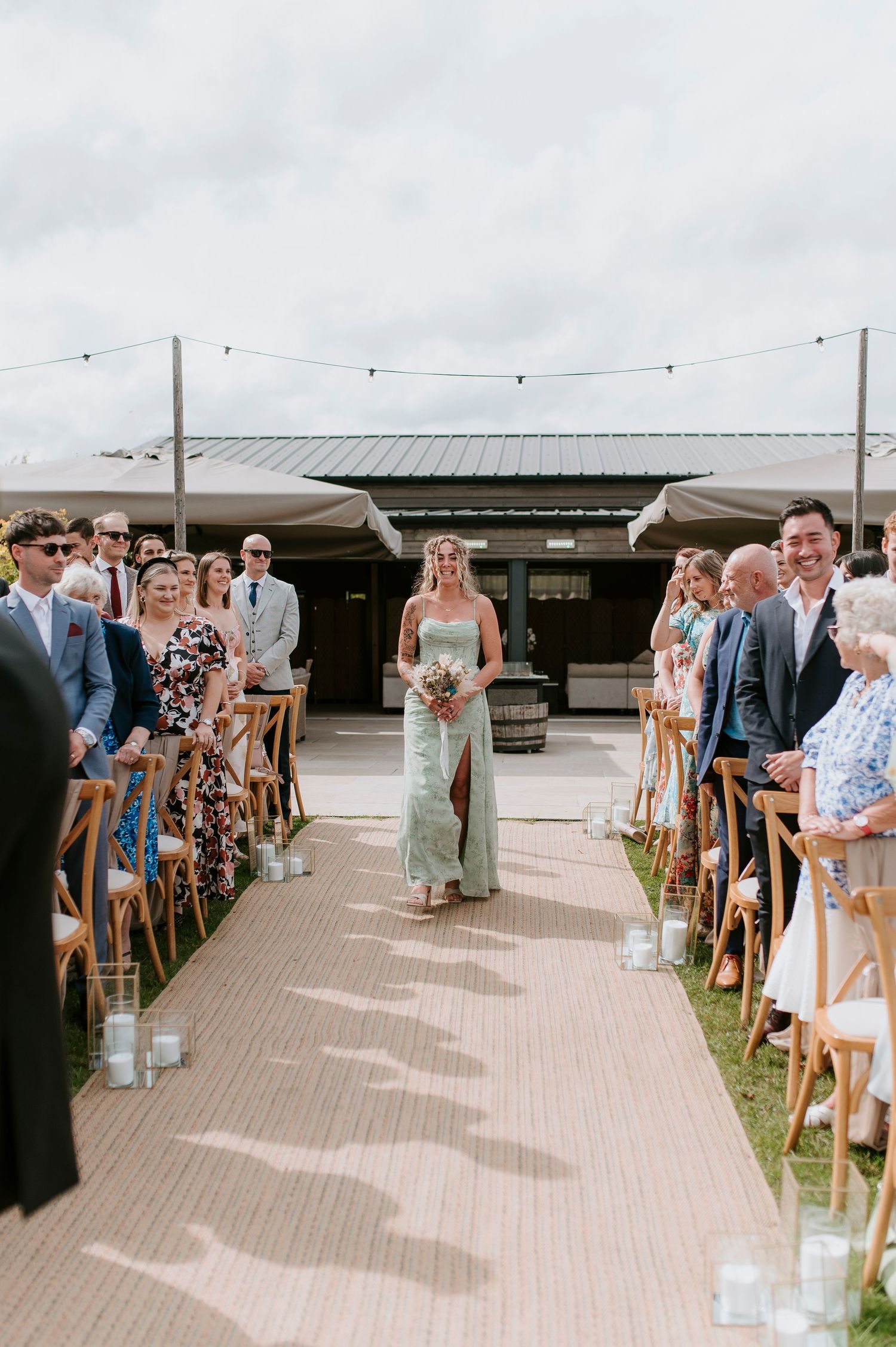 Wedding ceremony setup with wooden chairs lining an outdoor aisle decorated with hanging glass lanterns and flowers.