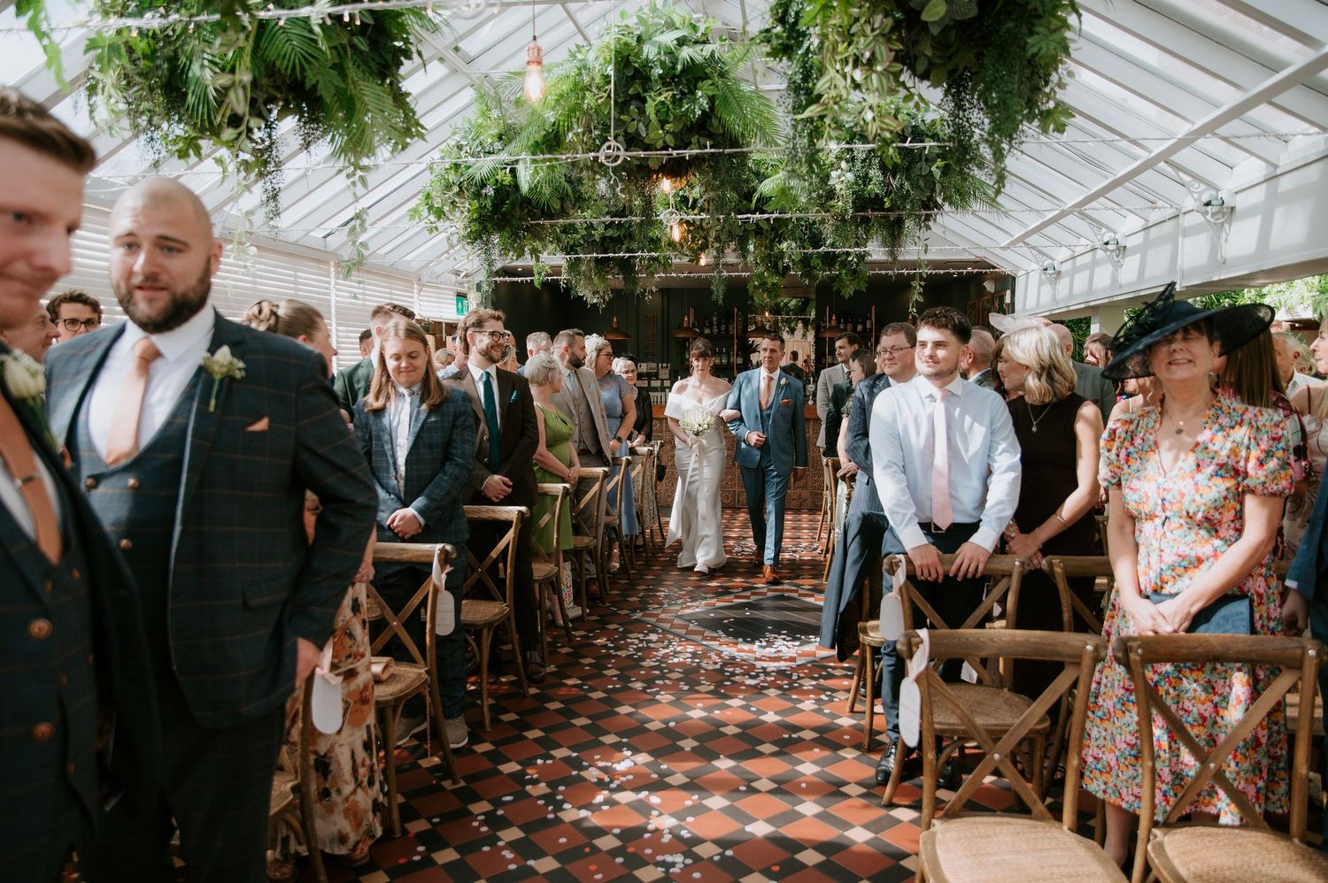Wedding ceremony taking place in a greenhouse venue with natural light streaming through glass panels and lush greenery overhead.