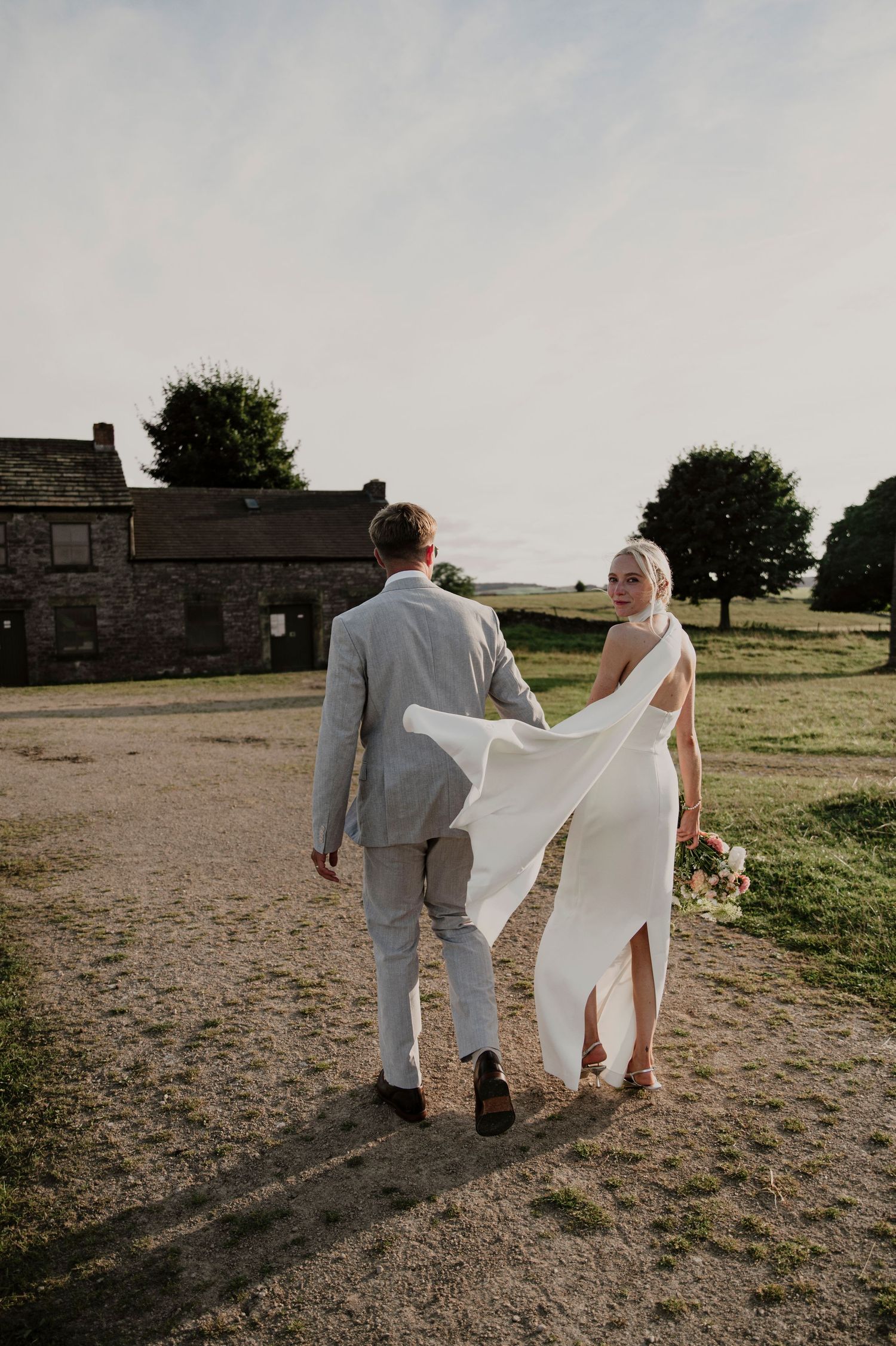 Newlywed couple walking together across a rustic field at sunset with a stone building in the background.