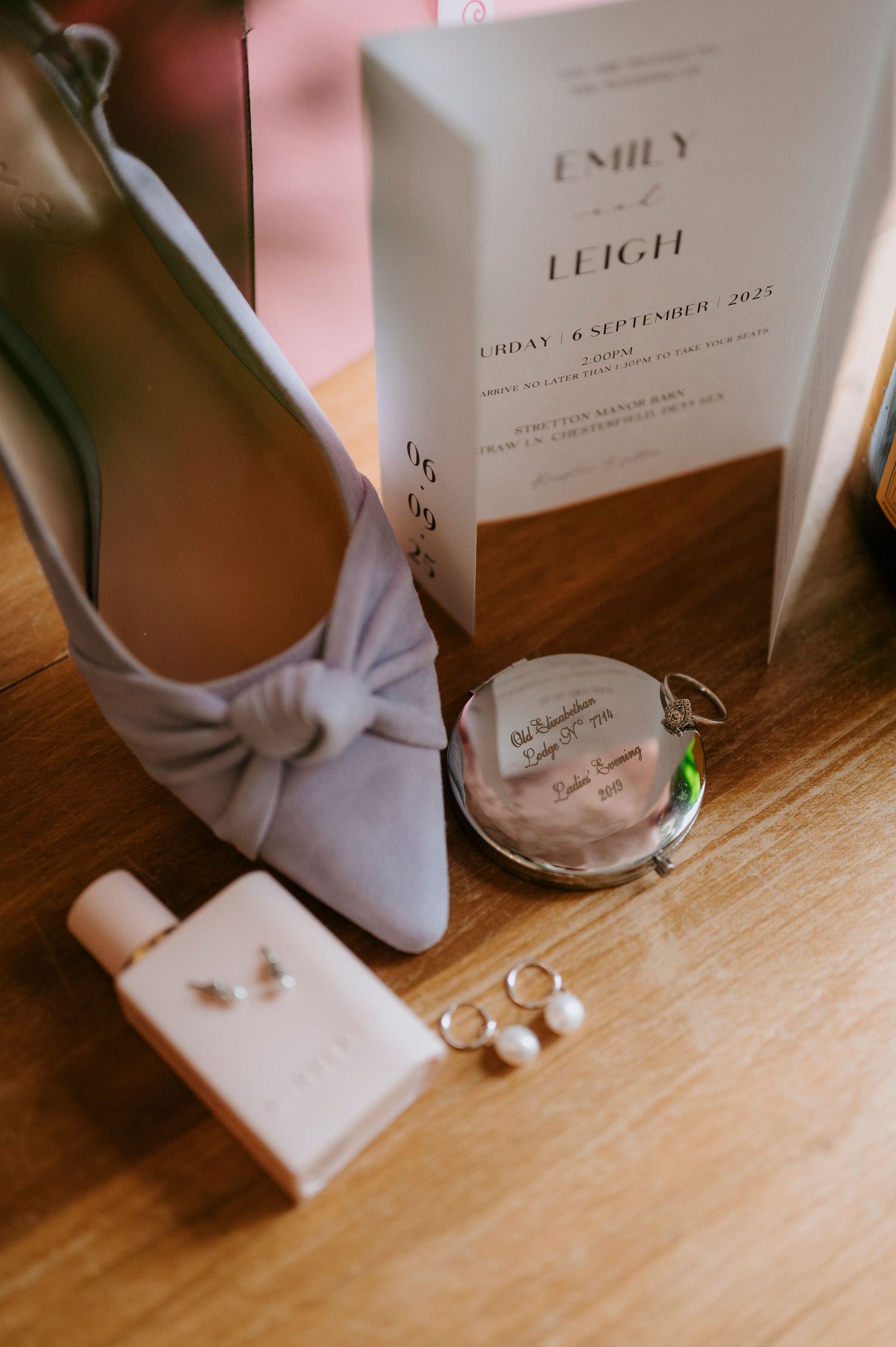 Wedding details including a gray bow heel, invitation, rings, and accessories arranged on a wooden surface.